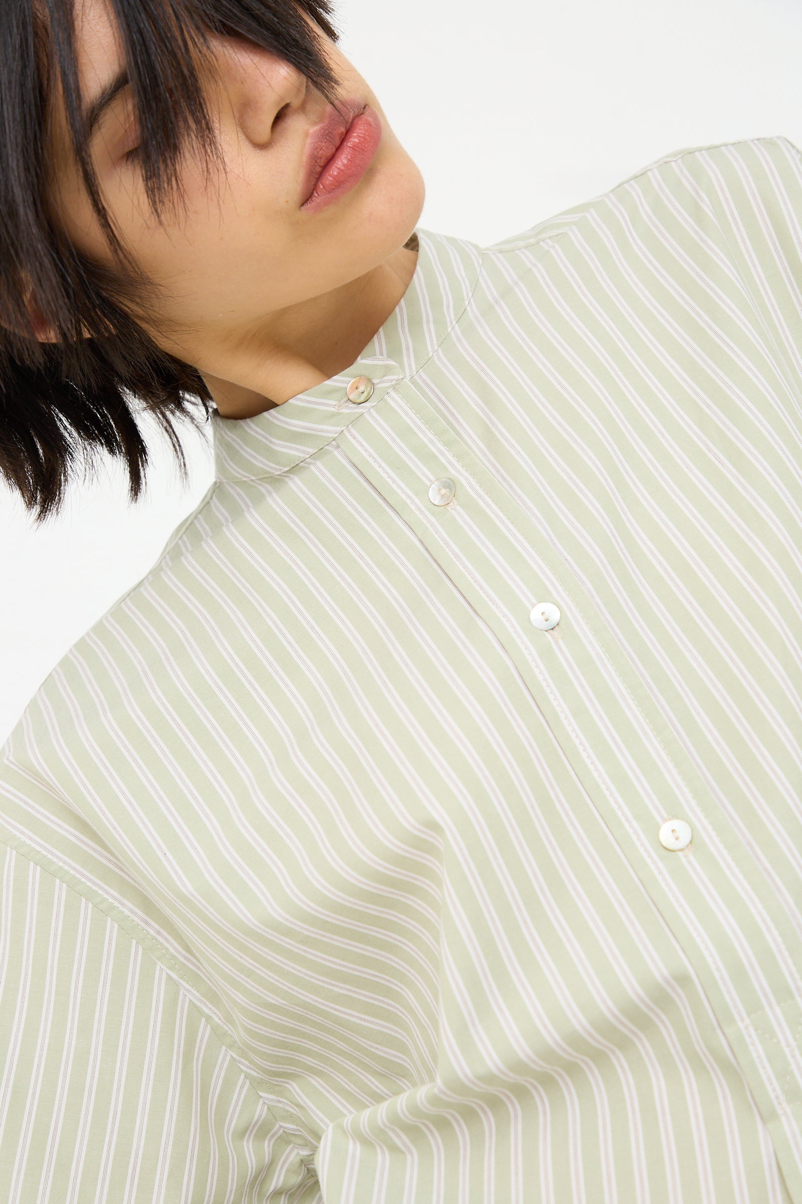 A close-up of a person in a Cawley Japanese Cotton Ines Shirt in Mint and Ecru with a buttoned collar. The person's face is partially obscured by their dark hair, and their eyes are closed.