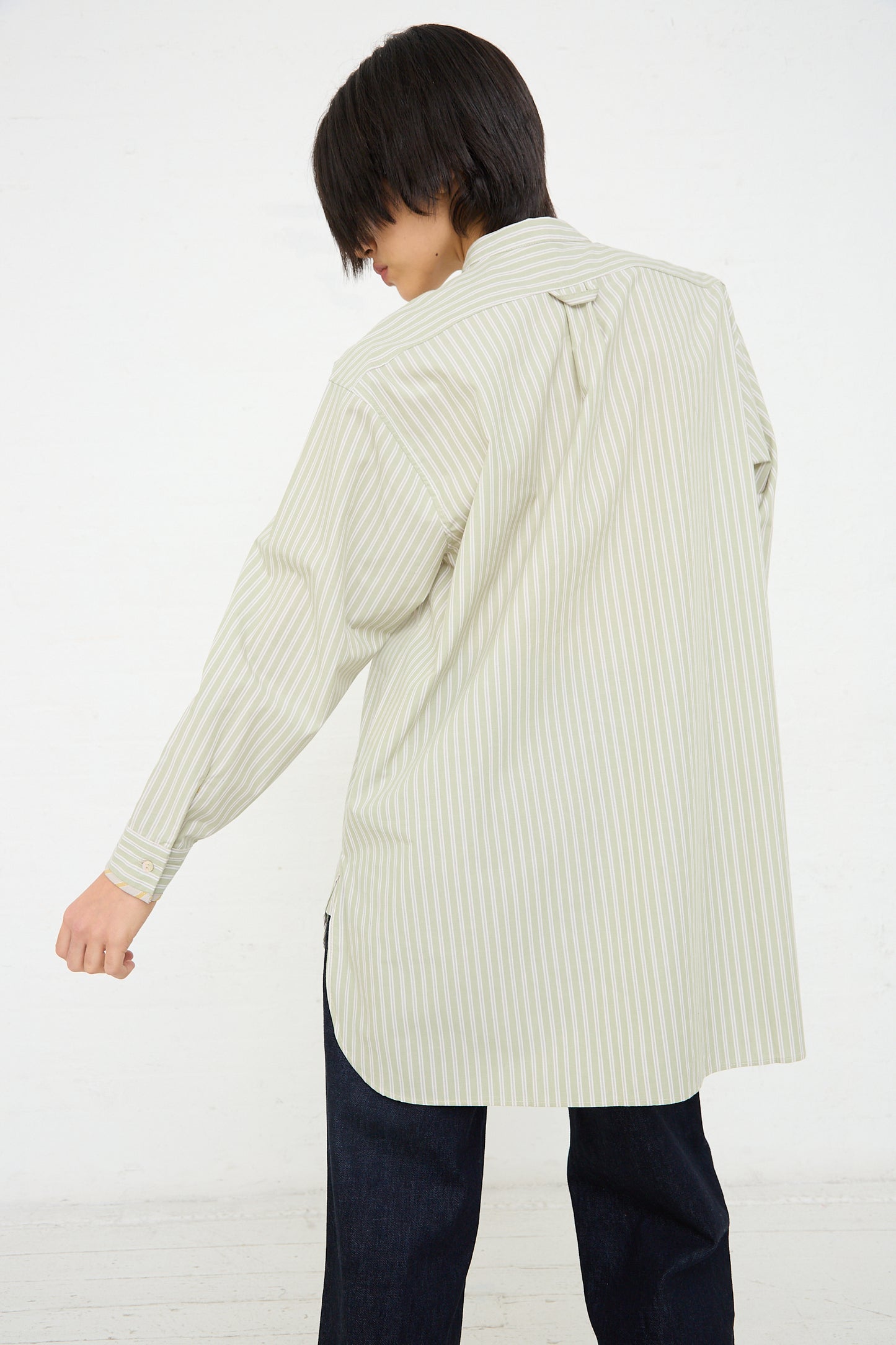 A person is seen from behind wearing a striped button-up shirt, crafted from British-manufactured cotton, with a vertical green and white pattern, standing against a white background. The shirt is the Japanese Cotton Ines Shirt in Mint and Ecru by Cawley.