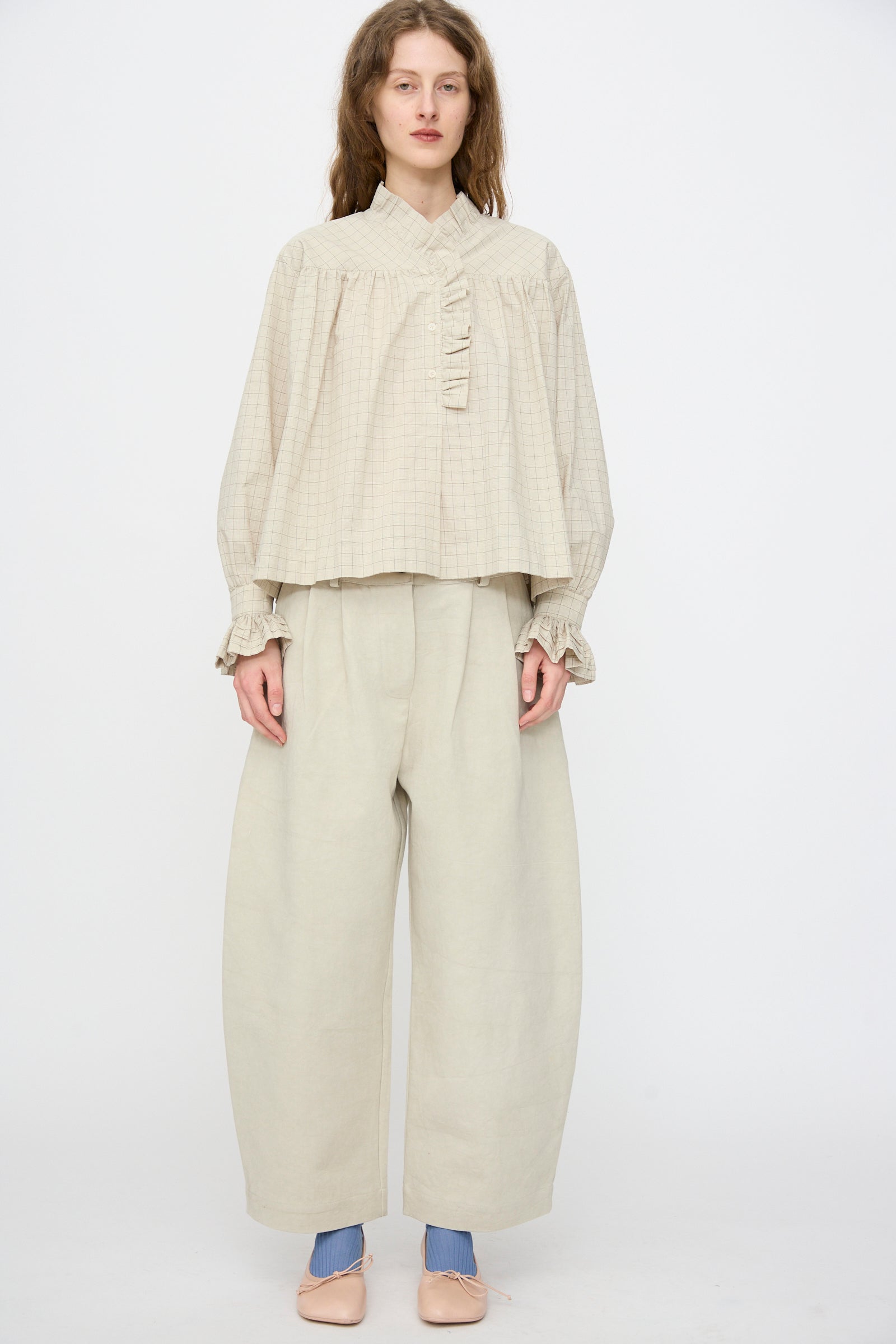 A person stands against a plain background in a loose, beige checked top with ruffled trim and Cawley Japanese Cotton Linen Robyn Trouser in Natural, styled with pale pink shoes.
