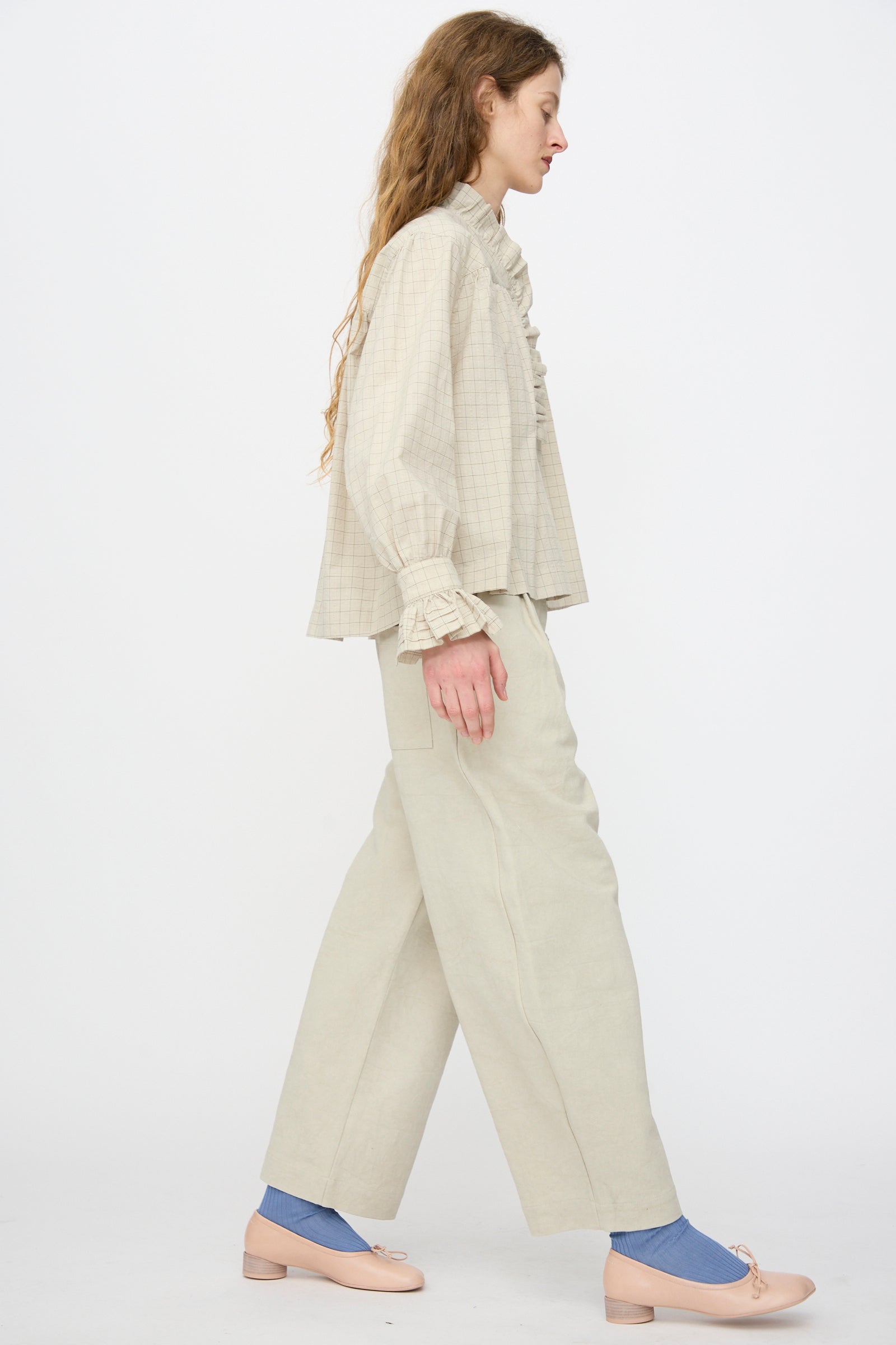 A woman with long, wavy hair walks in profile, wearing a loose beige checked blouse, Cawley’s Japanese Cotton Linen Robyn Trouser in Natural with a balloon leg silhouette, blue socks, and nude flats against a plain white background.