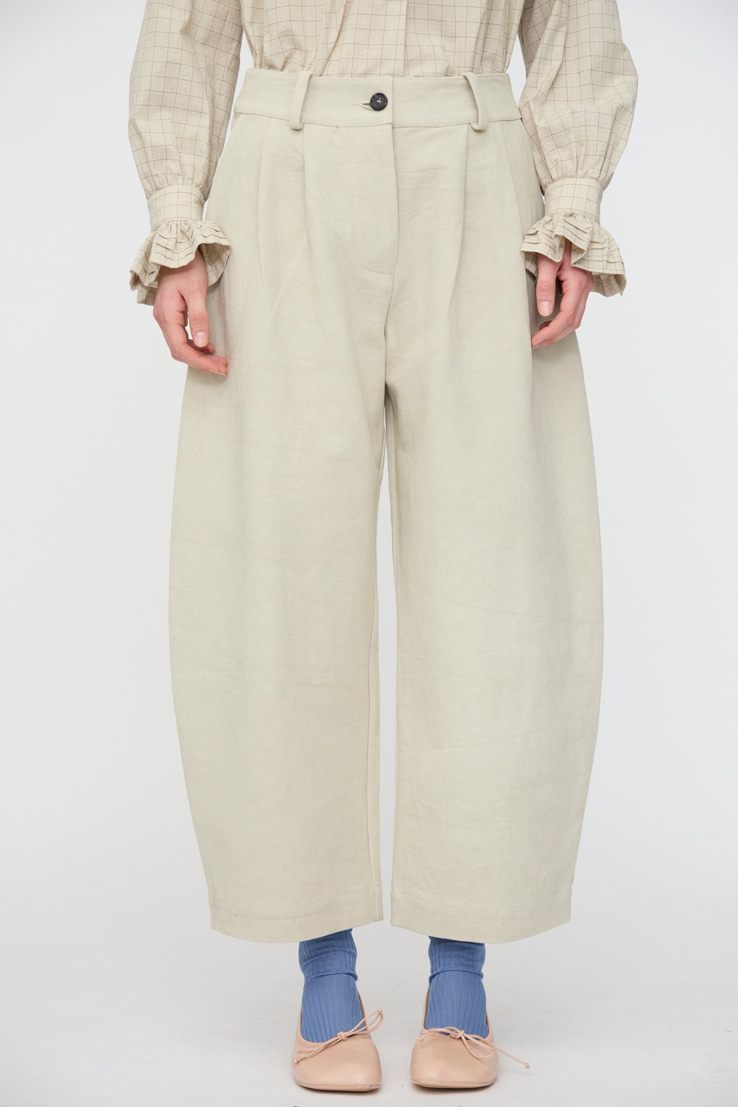 A person stands against a plain white background wearing Cawley’s Japanese Cotton Linen Robyn Trouser in Natural, paired with a beige plaid blouse with ruffled cuffs, blue socks, and beige flat shoes.