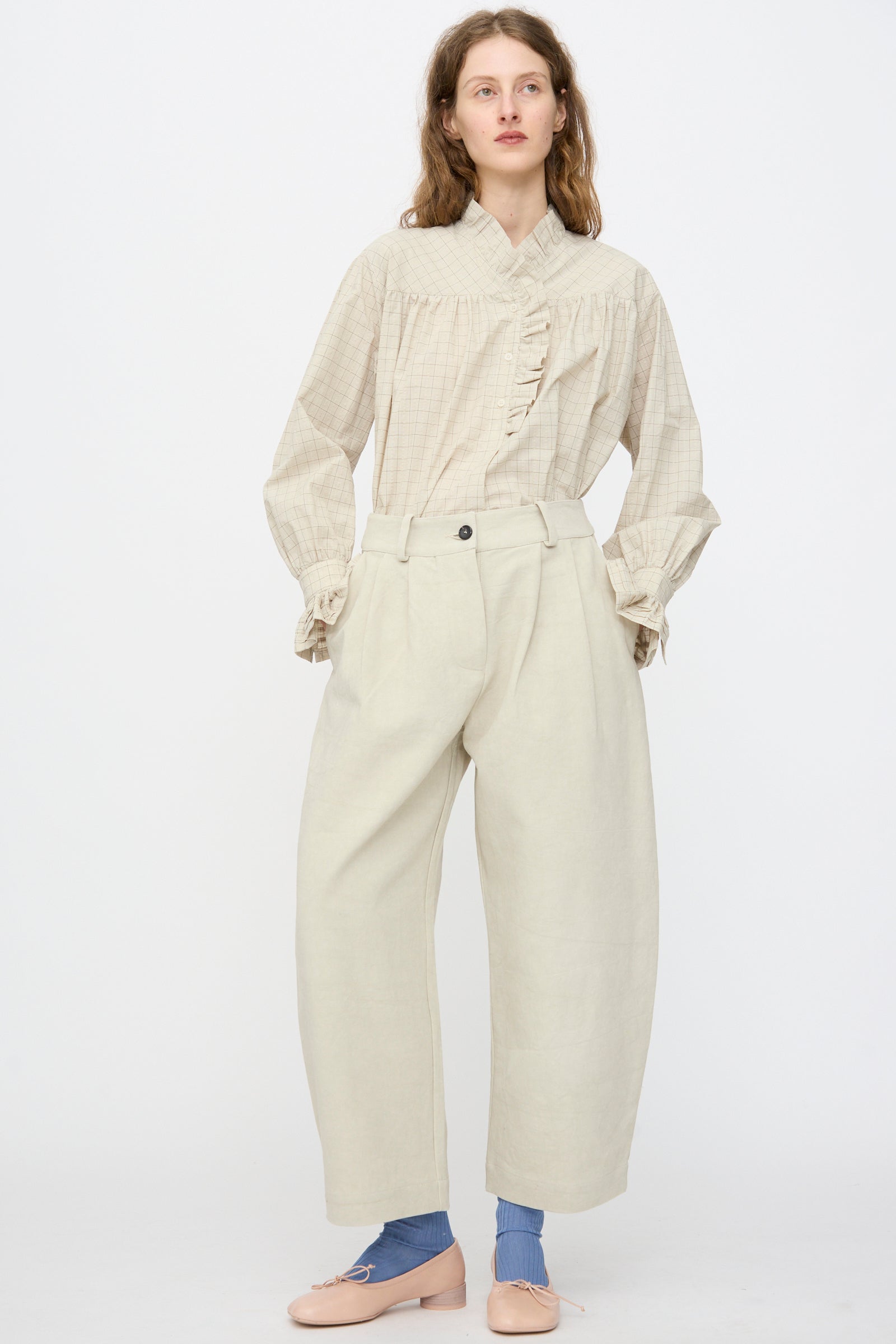 A person stands against a white background wearing a beige long-sleeve blouse with ruffles, Cawley’s Japanese Cotton Linen Robyn Trouser in Natural in a balloon leg silhouette, blue socks, and pale pink flats.