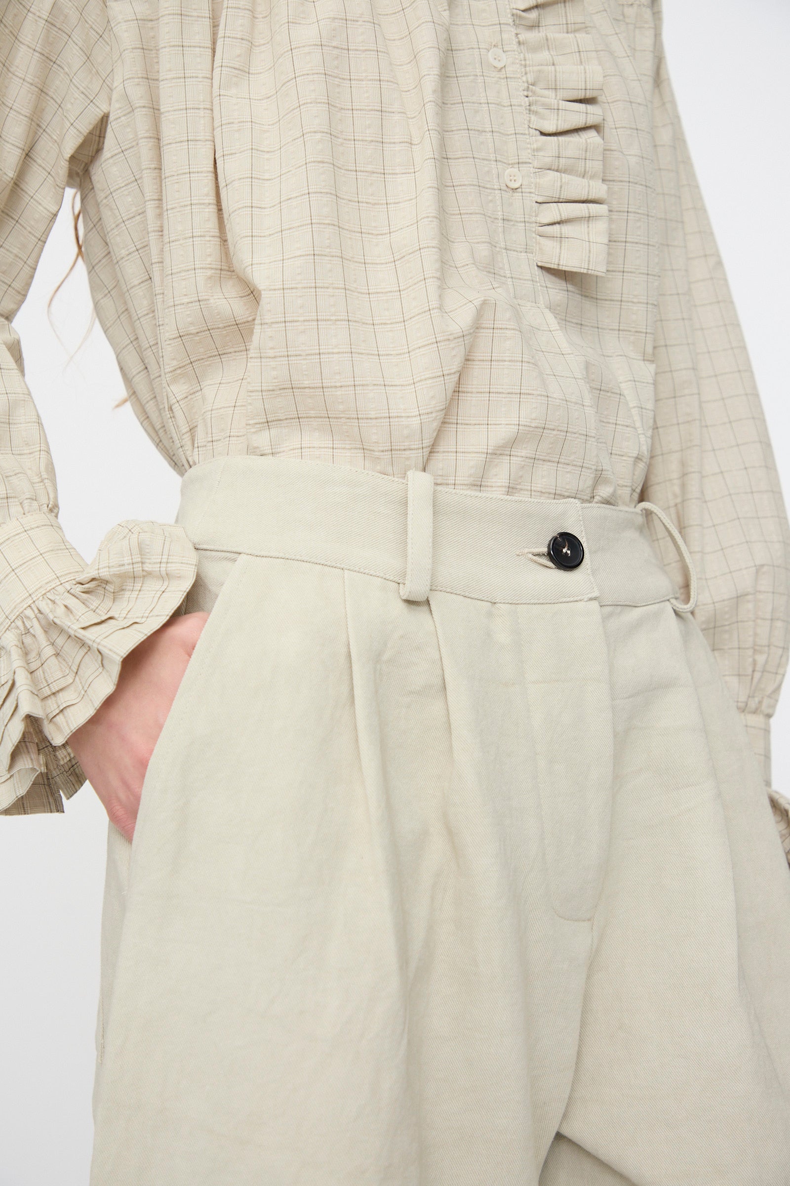 A person wears a beige checkered ruffle long-sleeve blouse and Cawley’s Japanese Cotton Linen Robyn Trouser in Natural with a balloon leg silhouette, one hand in the pocket.