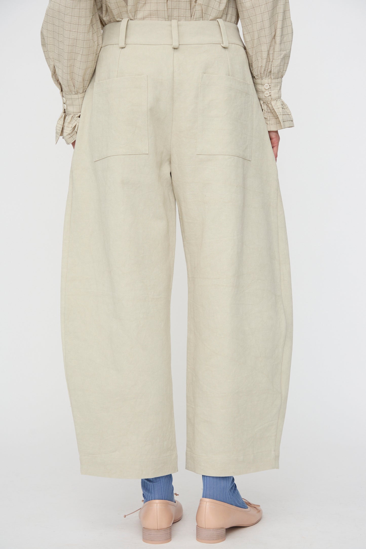 A person is seen from the back in Cawley’s Japanese Cotton Linen Robyn Trouser in Natural—high-waisted with patch pockets, balloon leg silhouette, made in London—styled with a checked blouse and blue socks.