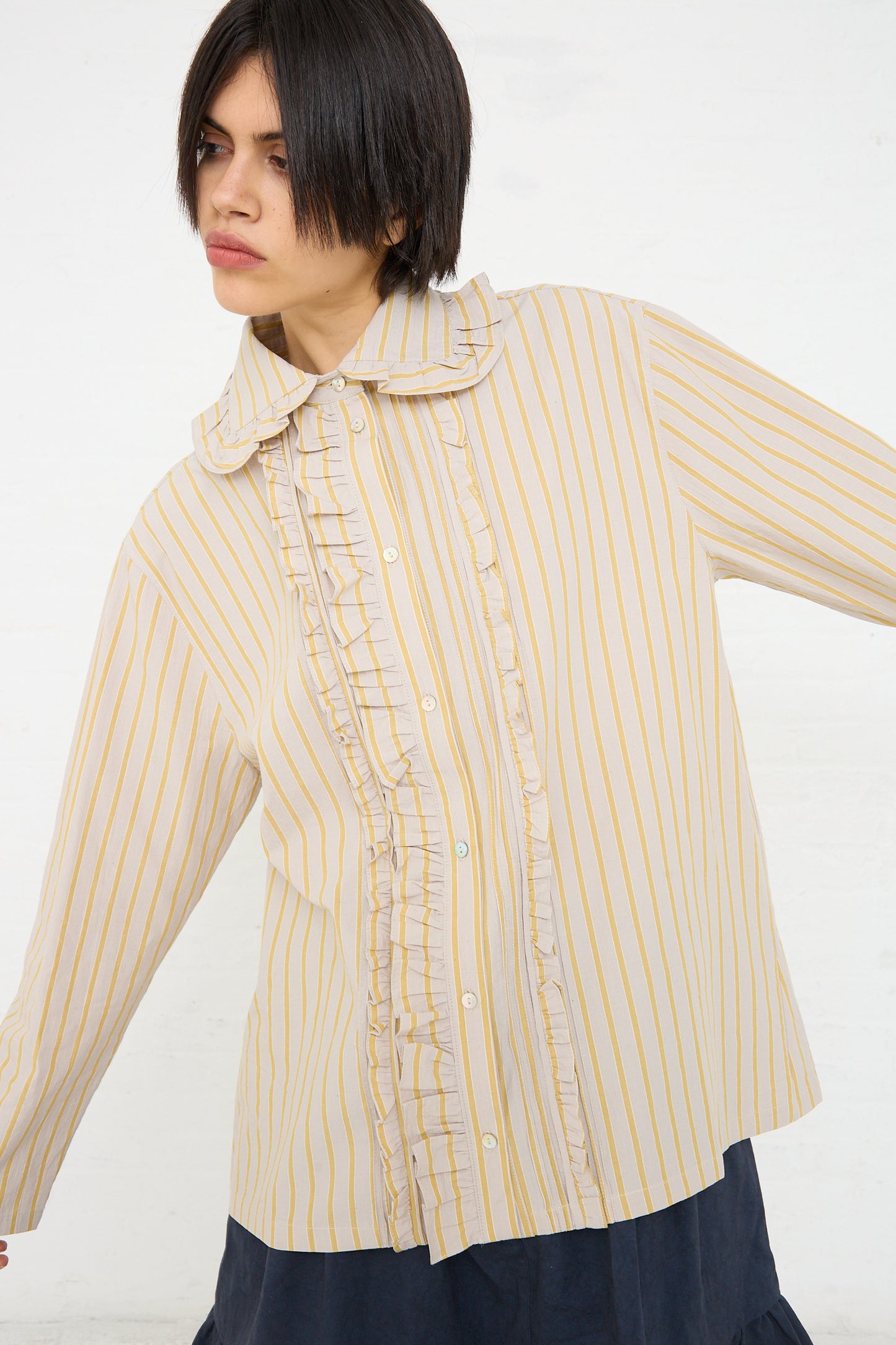 A woman with short black hair wearing a Cawley Japanese Cotton Ruffle Shirt in Sun and Grey, looking to the side with a slight frown on her face.