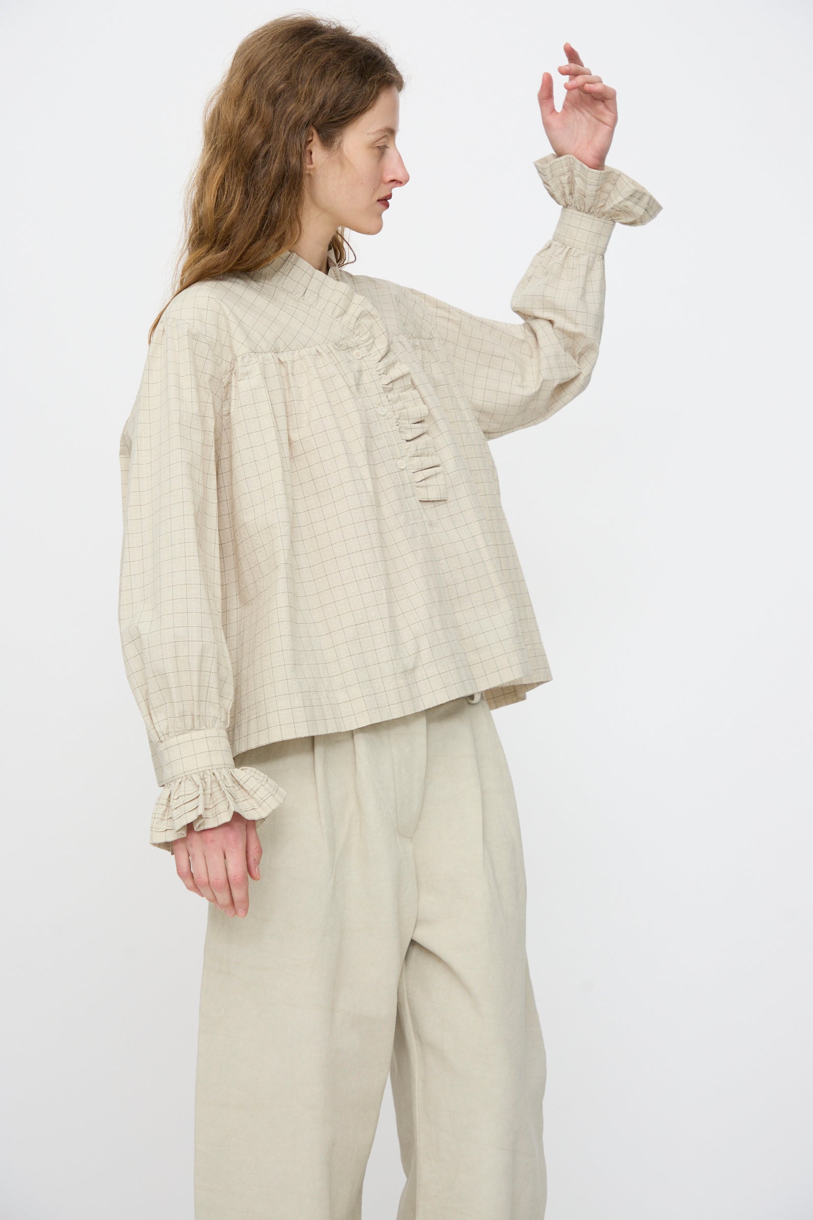 A person wearing the Cawley Japanese Cotton Sophie Shirt in Natural Check, featuring a ruffle collar, and wide-leg pants, stands against a plain white background.
