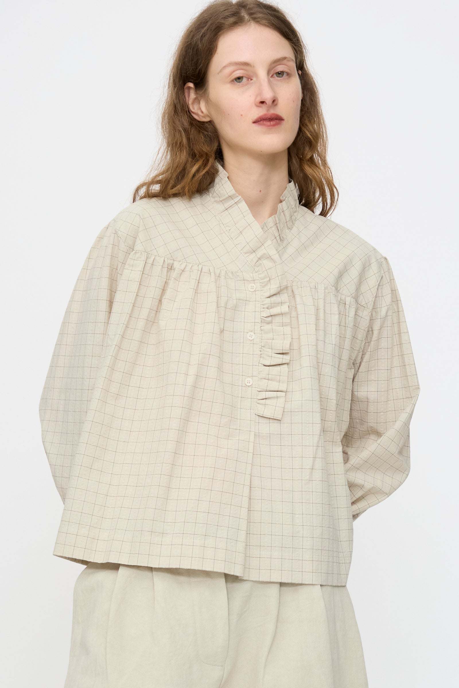 A person with long light brown hair wears the Cawley Japanese Cotton Sophie Shirt in Natural Check, featuring a ruffle collar and buttons, standing against a plain background.