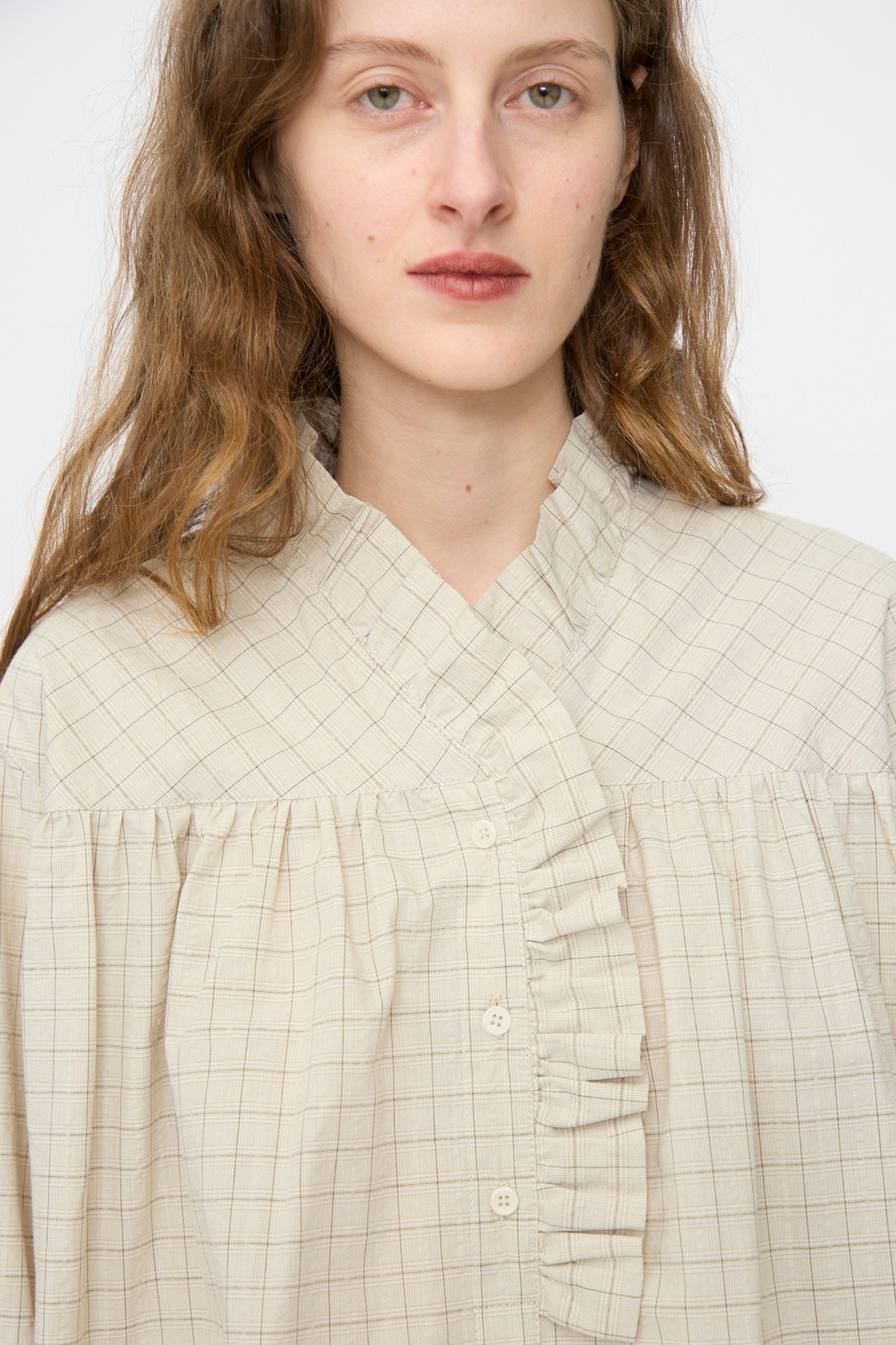 A person with long brown hair models the Cawley Japanese Cotton Sophie Shirt in Natural Check, featuring ruffle details and a high collar, looking directly at the camera against a plain background.