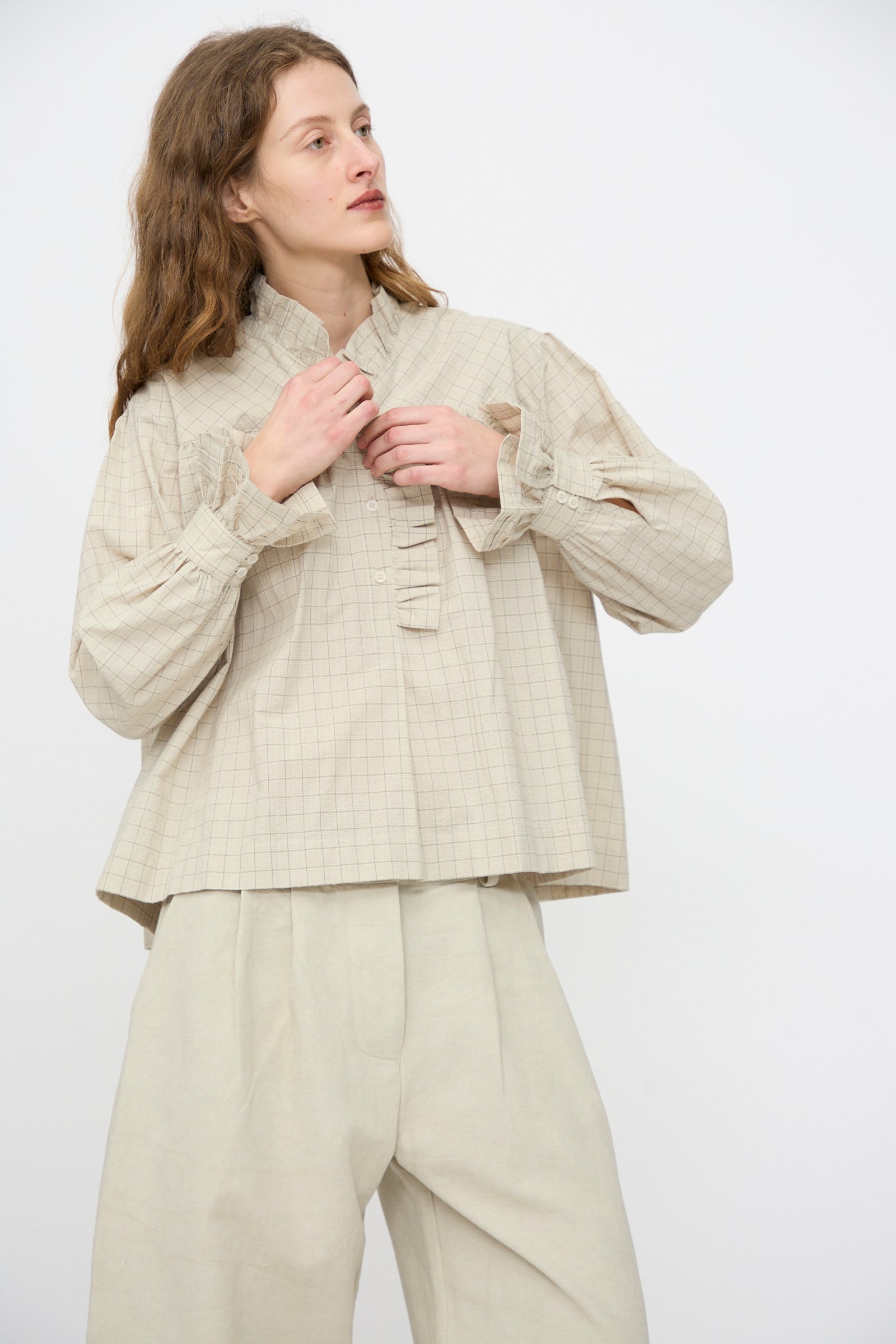 A person wears the Cawley Japanese Cotton Sophie Shirt in Natural Check, featuring a subtle ruffle collar, paired with wide-legged beige pants, standing against a plain white background.