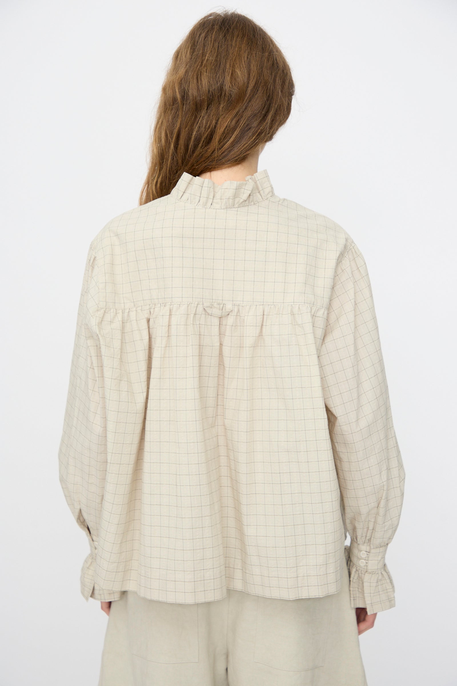 A person with long brown hair is seen from behind, wearing the Cawley Japanese Cotton Sophie Shirt in Natural Check—a loose-fitting blouse with a plaid pattern, ruffle collar, and cuffs.