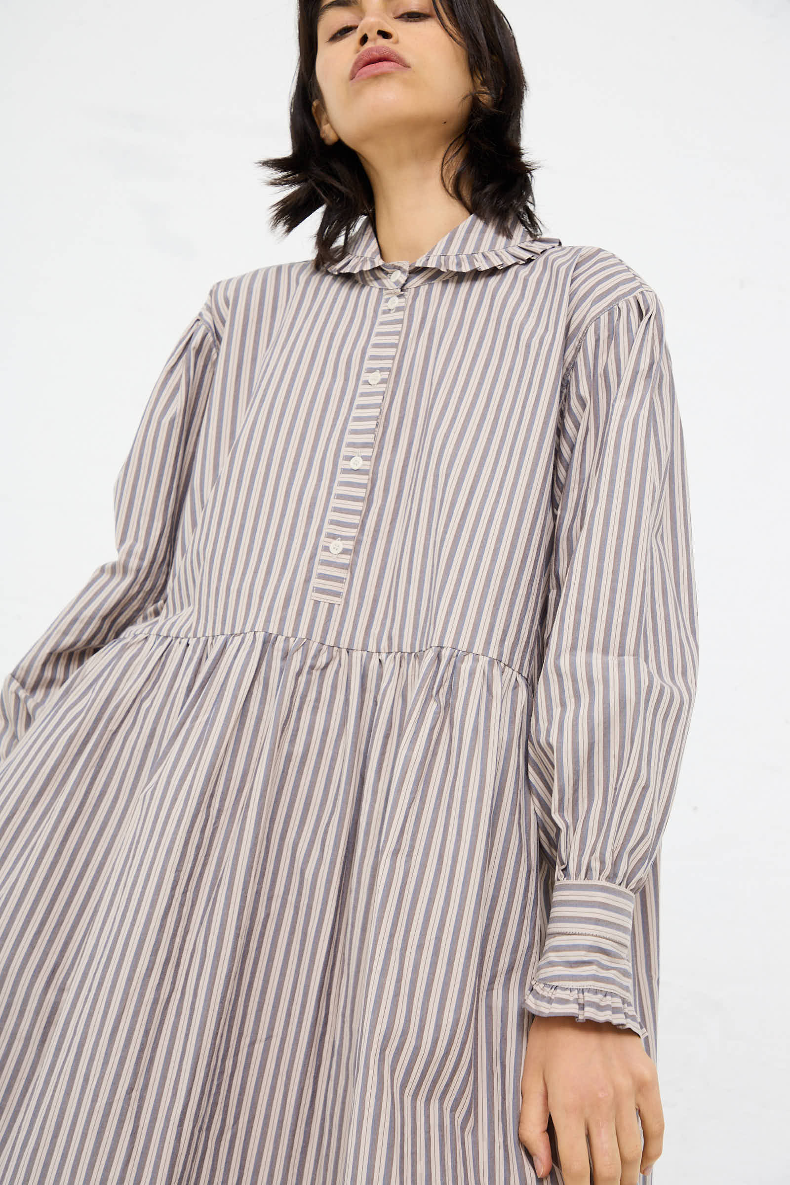 A person wearing the Lydbrook Dress in Natural, Blue and Brown Stripe by Cawley, an oversized, long-sleeved Japanese cotton dress with a collar, looking slightly upward.