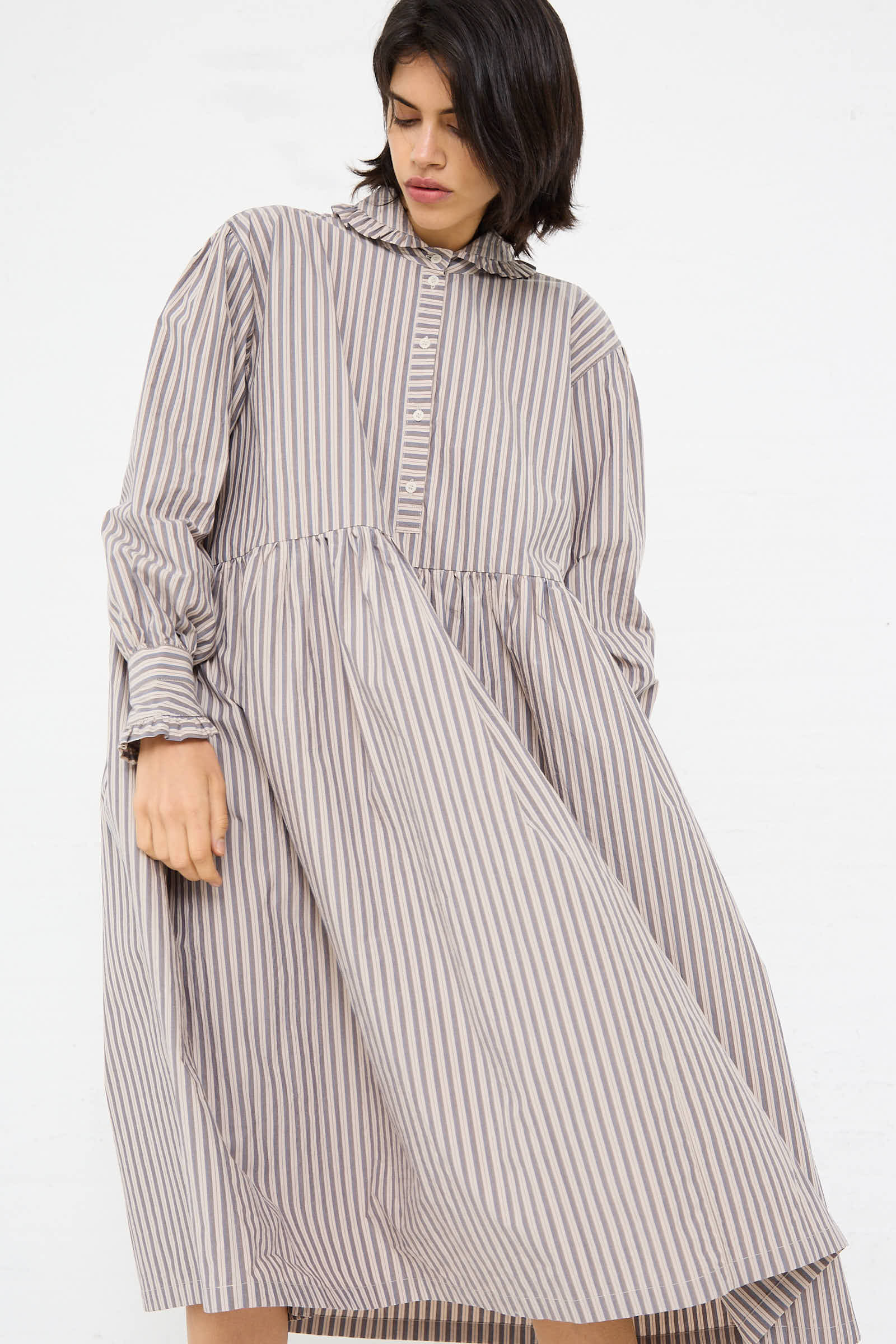 A person wearing the Cawley Lydbrook Dress in Natural, Blue and Brown Stripe, a long-sleeved, loose-fitting midi dress made of Japanese striped cotton with a ruffled neck collar, is looking downwards.