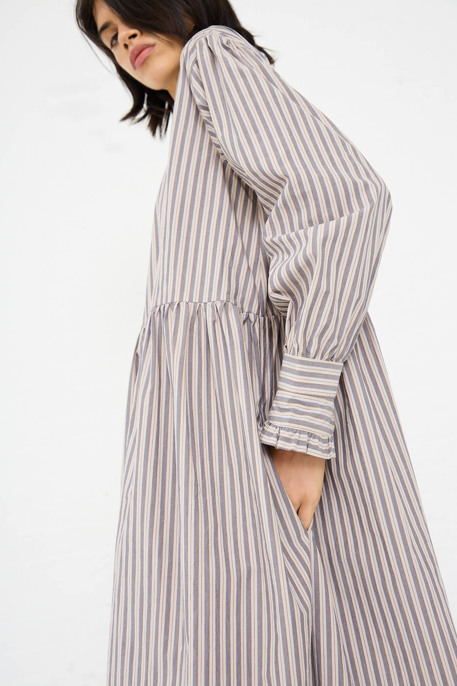 A person wearing the Lydbrook Dress in Natural, Blue and Brown Stripe by Cawley, a loose-fitting, long-sleeve, Japanese striped cotton midi dress with hands in pockets looks slightly downward.