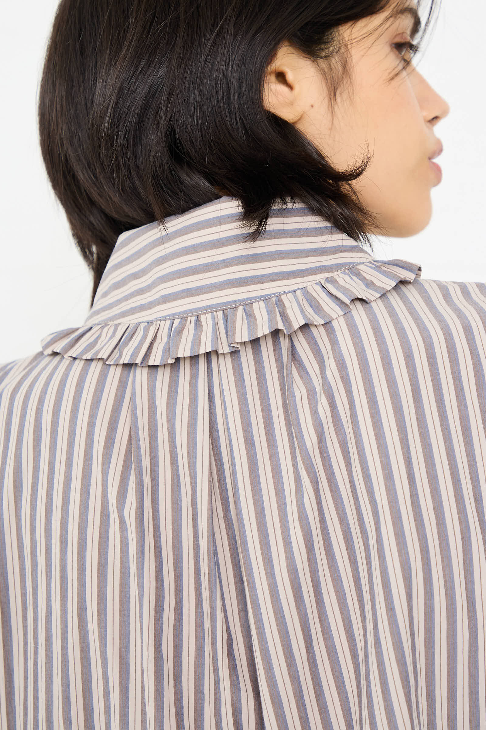 A person with short black hair, viewed from behind, is wearing the Lydbrook Dress in Natural, Blue and Brown Stripe by Cawley, featuring a ruffled collar and made from Japanese striped cotton.