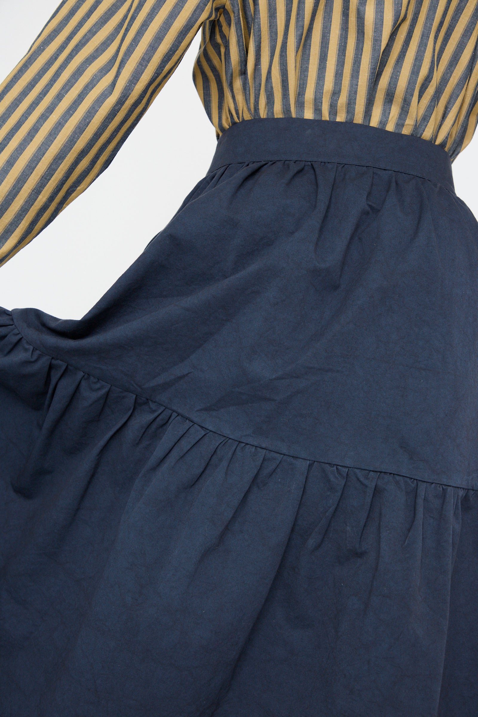 Close-up of a person wearing a striped yellow and blue blouse paired with the Cawley Japanese Cotton Twill Patience Skirt in Dark Navy, against a plain backdrop. 