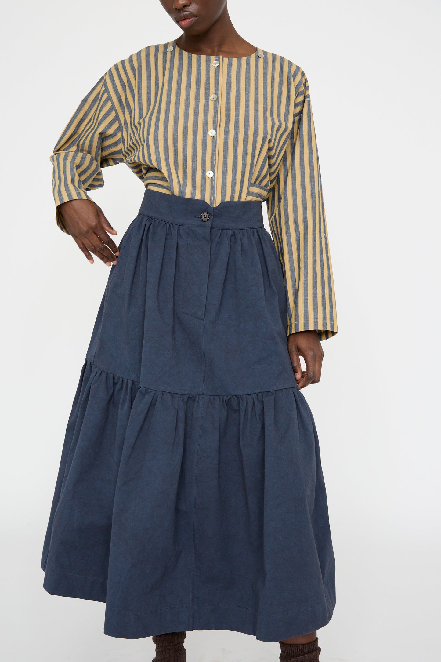 A person stands against a plain white background wearing a striped button-up shirt and the Cawley Japanese Cotton Twill Patience Skirt in dark navy.