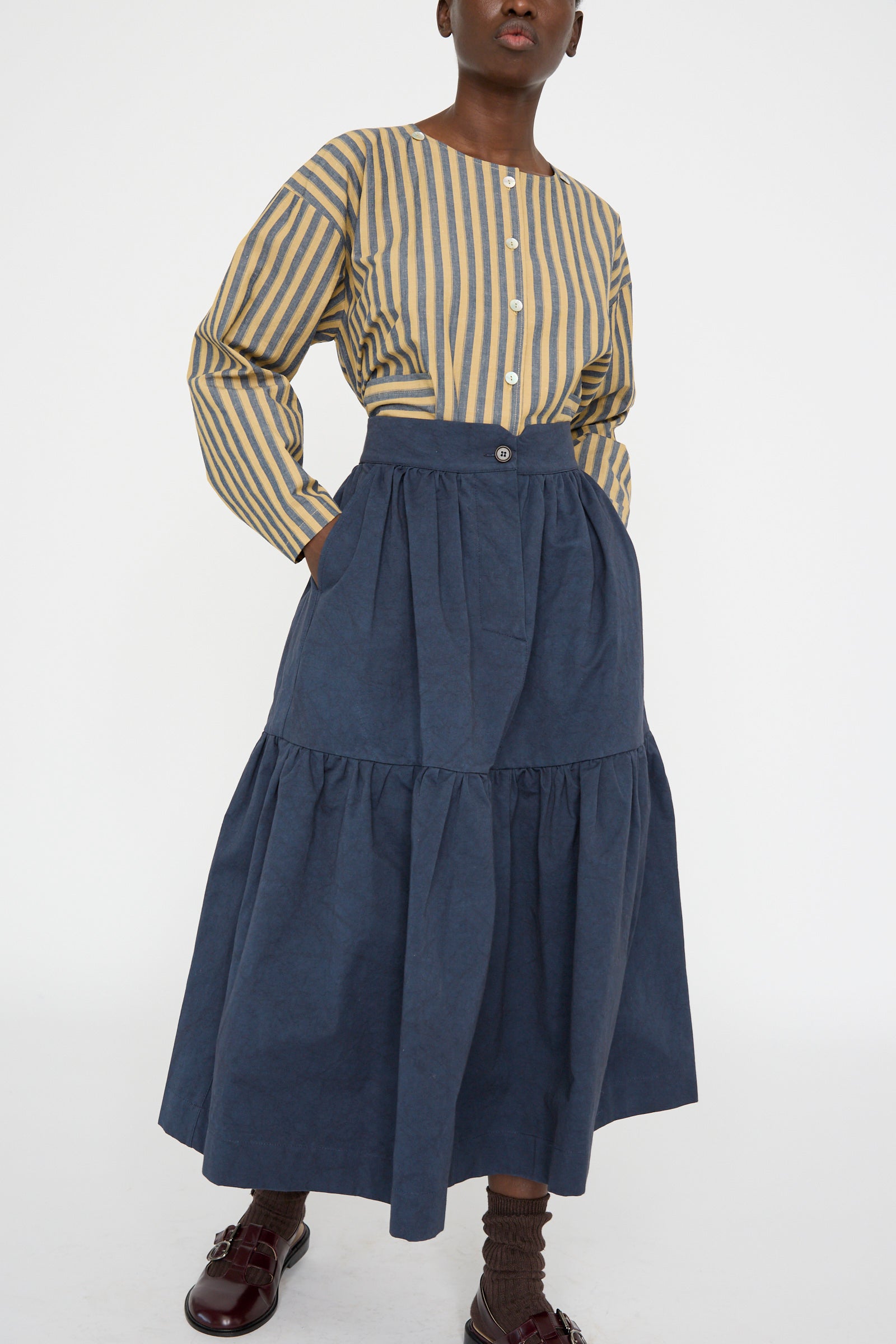 A person in a striped long-sleeve shirt and Cawley's Japanese Cotton Twill Patience Skirt in Dark Navy stands against a plain backgroundm with hands in their pockets.