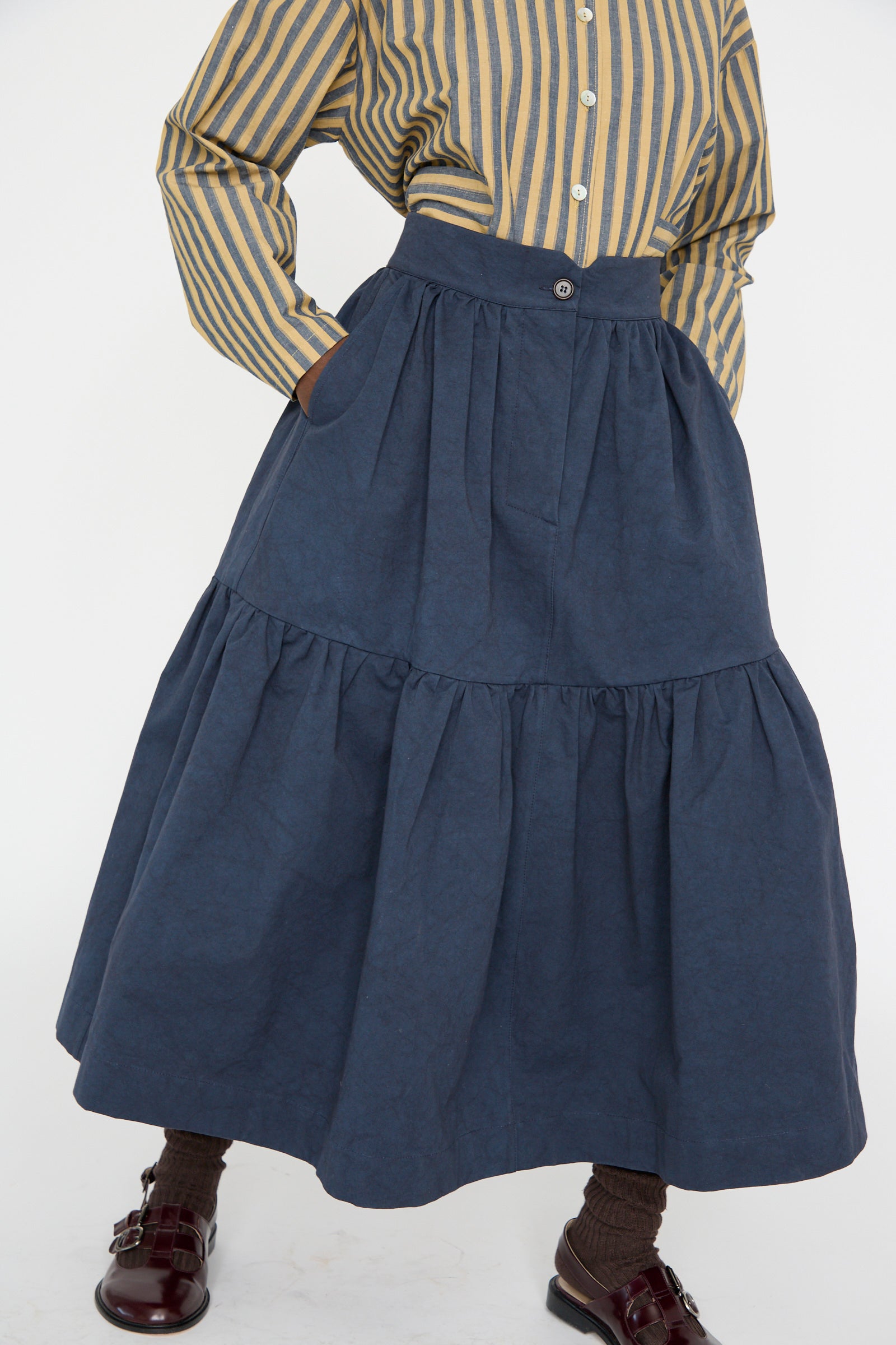 The model is wearing a yellow and black striped shirt with the Cawley Japanese Cotton Twill Patience Skirt in Dark Navy, brown socks, and burgundy shoes, seen from below the shoulders. 