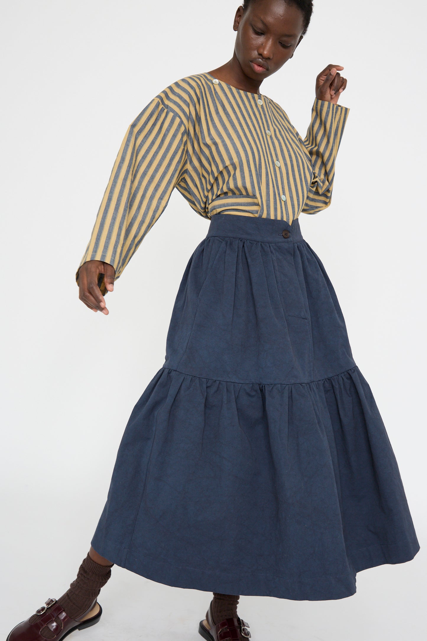 A person wearing a striped shirt and the Cawley Japanese Cotton Twill Patience Skirt in Dark Navy poses against a plain background.