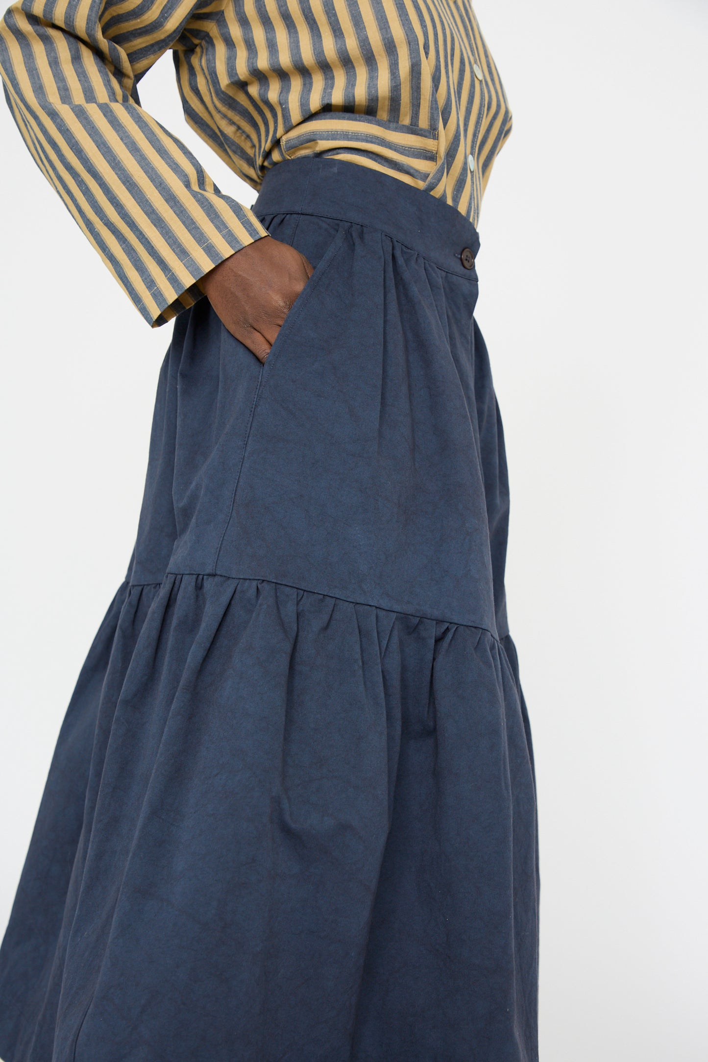 A close up of a person wearing the Cawley Japanese Cotton Twill Patience Skirt in Dark Navy paired with a striped yellow and gray top, standing with one hand in their pocket.