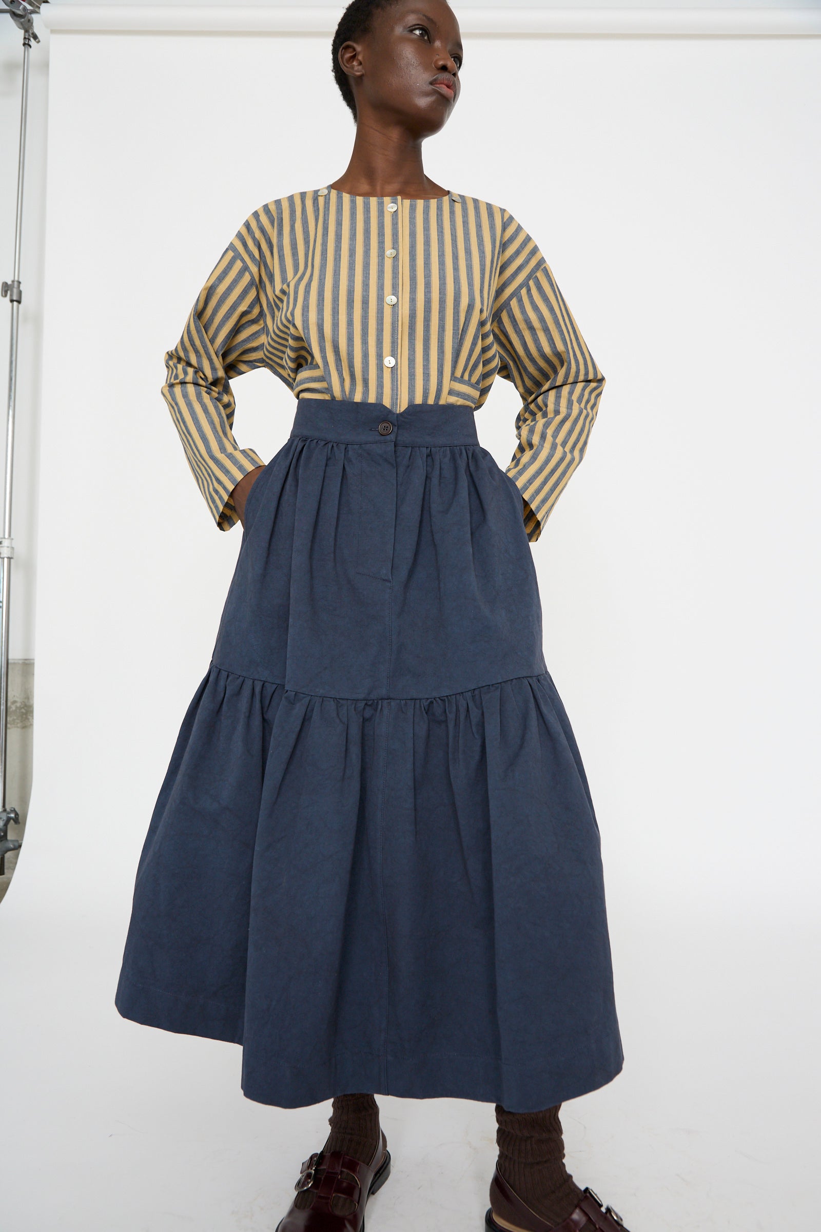 A person wearing a striped long-sleeve shirt and the Cawley Japanese Cotton Twill Patience Skirt in Dark Navy stands against a white backdrop.