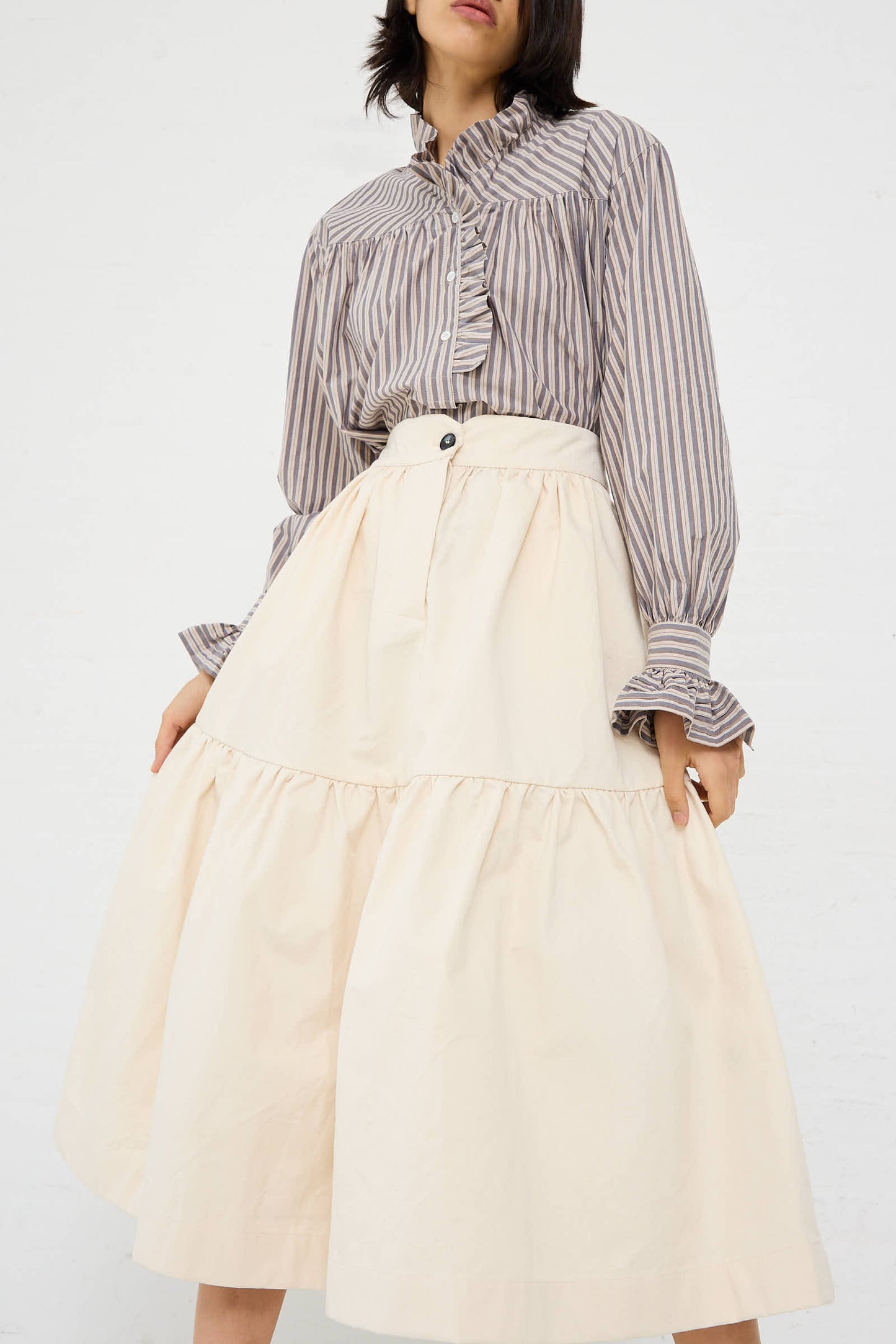 A person wearing a loose-fitting, striped blouse and the Cawley Japanese Cotton Twill Patience Skirt in Ecru stands against a plain white background.