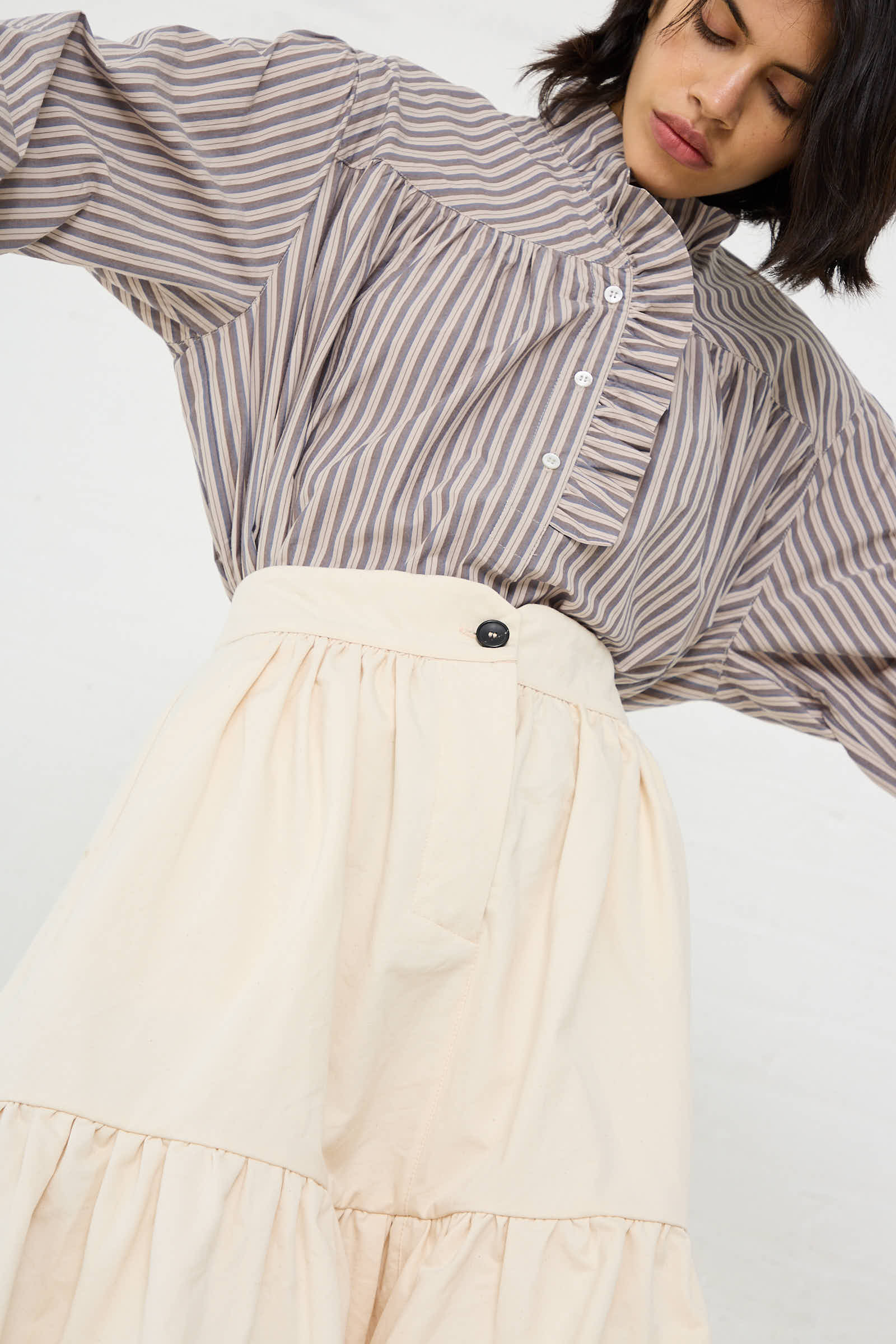 A woman is wearing a vertically striped button-up shirt with an exaggerated collar and the Cawley Japanese Cotton Twill Patience Skirt in Ecru. She is facing downward, tilting her head to the left.