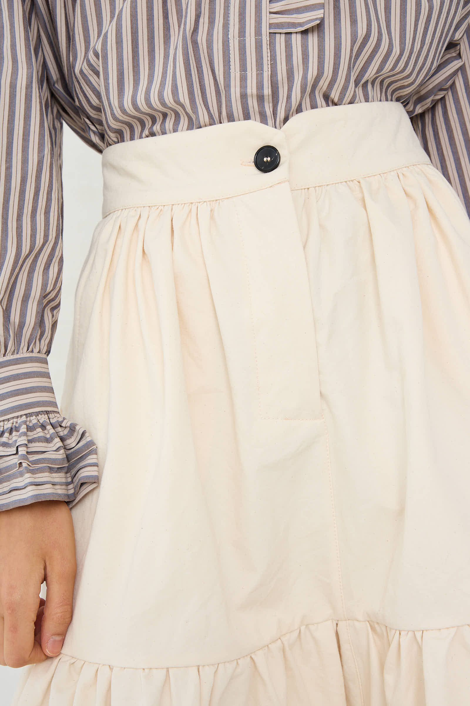 Individual dressed in a striped long-sleeve shirt with ruffled cuffs and the Cawley's Japanese Cotton Twill Patience Skirt in Ecru, which features a black button.