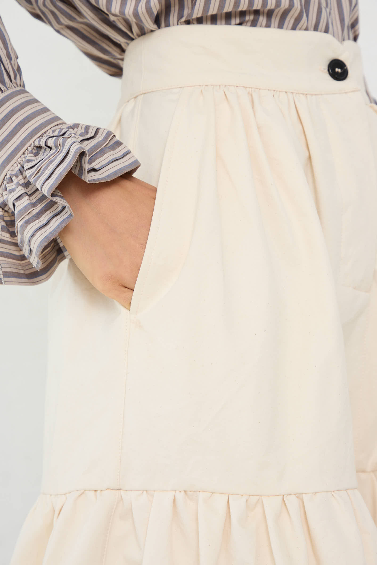 A person wearing a striped shirt with ruffled cuffs places their hand in the pocket of the Cawley Japanese Cotton Twill Patience Skirt in Ecru, which features a single black button at the waistband.