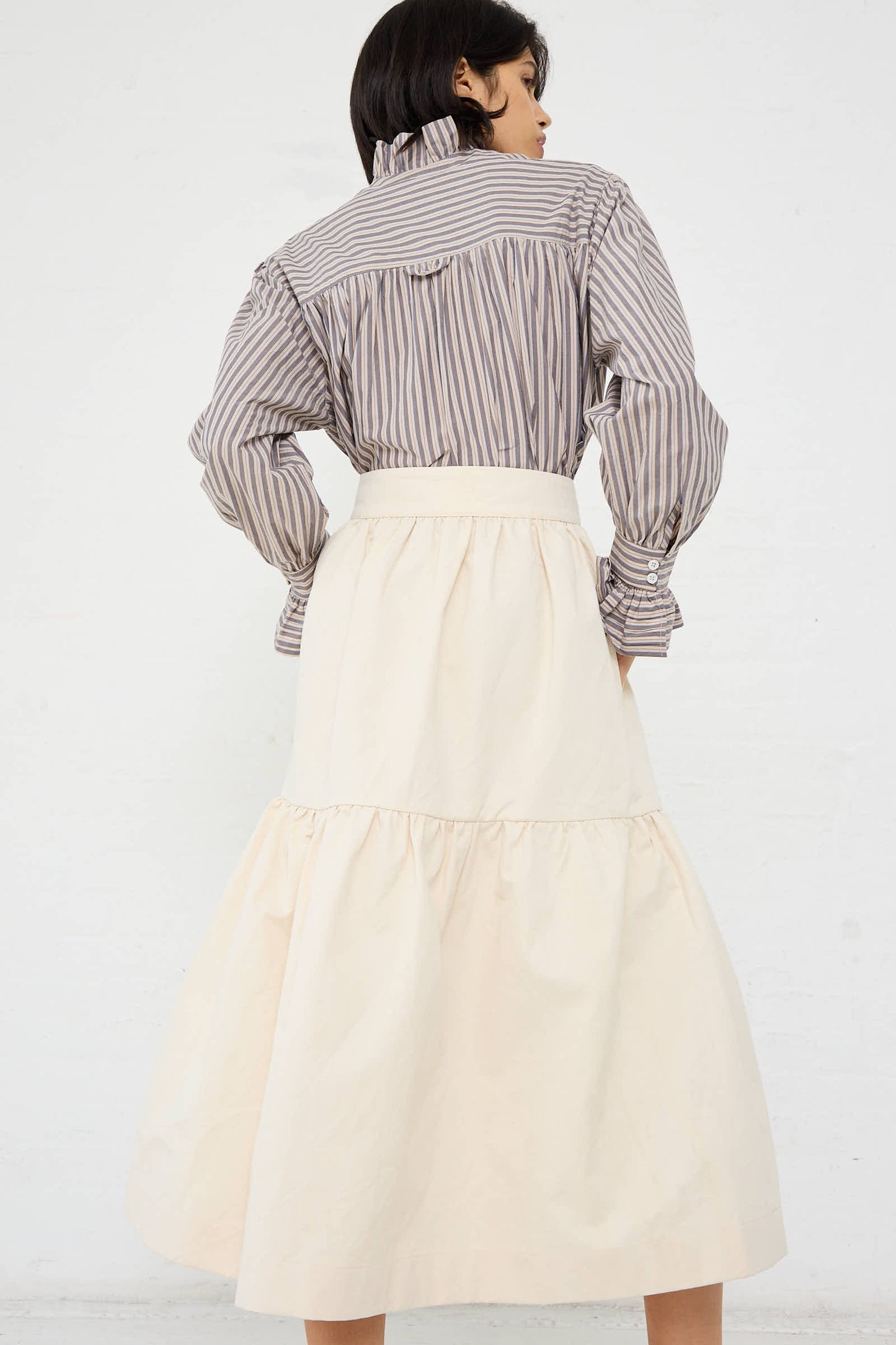 A person stands facing away, wearing a striped long-sleeve shirt with a high collar and the Cawley Japanese Cotton Twill Patience Skirt in Ecru.