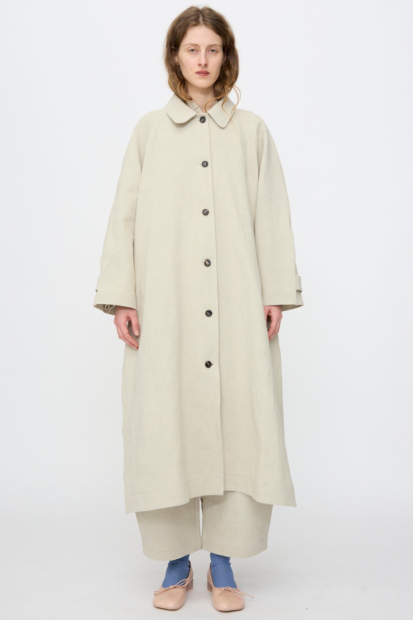A person stands facing forward against a plain white background, wearing the Cawley Japanese Drill Wickham Mac in Natural, wide beige pants, blue socks, and beige shoes.