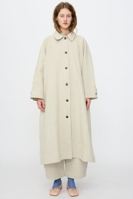 A person stands facing forward against a plain white background, wearing the Cawley Japanese Drill Wickham Mac in Natural, wide beige pants, blue socks, and beige shoes.