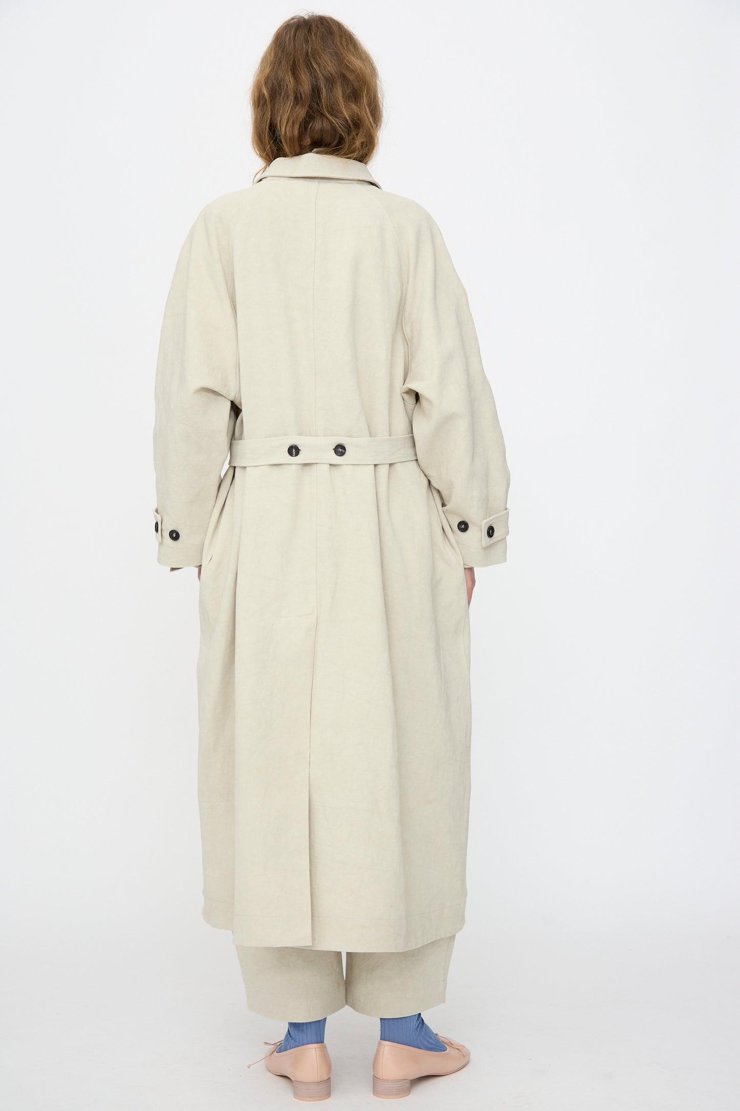 A person stands with their back to the camera, wearing Cawley's Japanese Drill Wickham Mac in Natural, paired with matching pants, pink shoes, and blue socks against a plain white background.