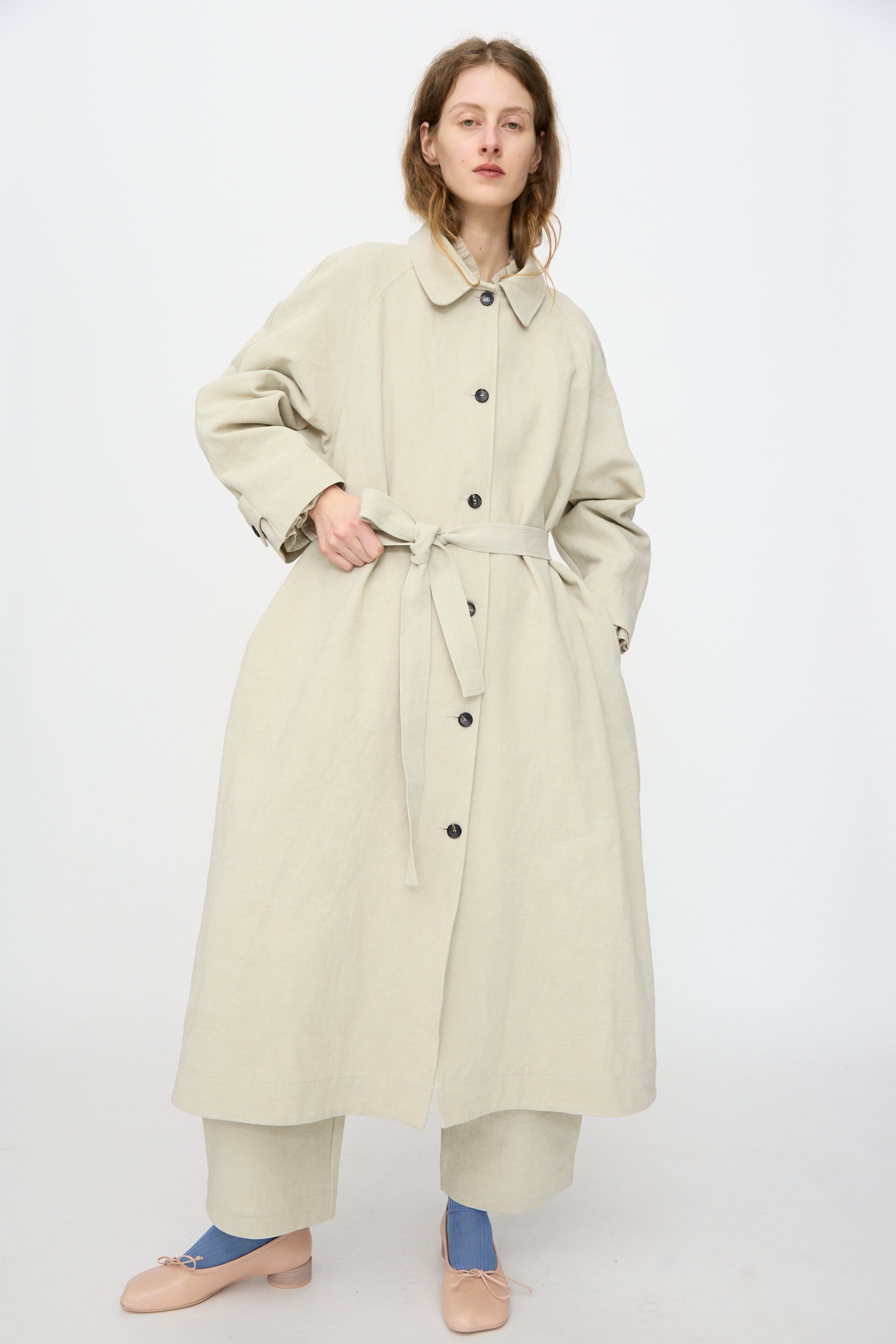 A person stands against a plain white background wearing the Cawley Japanese Drill Wickham Mac in Natural, styled with wide-leg pants and light pink shoes.