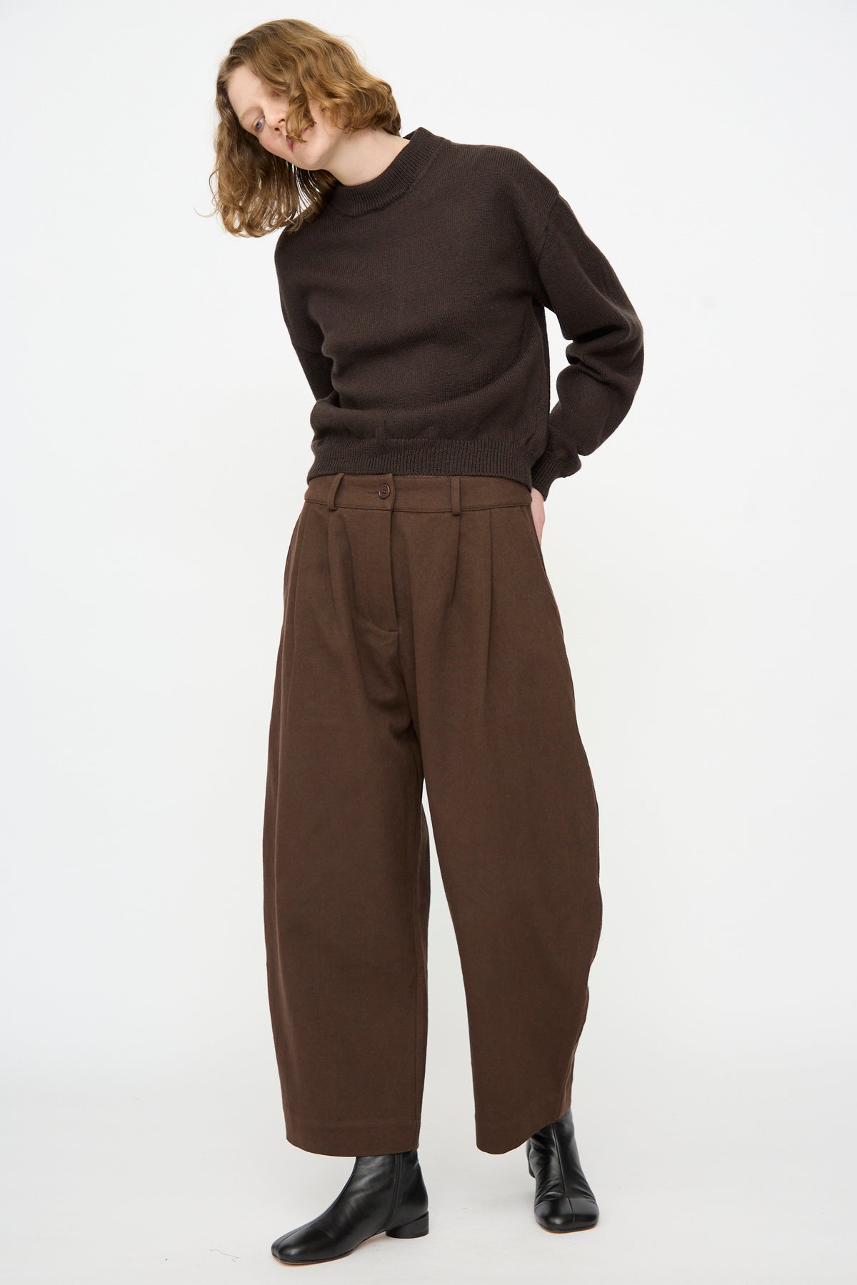 A person stands against a plain white background wearing a dark brown sweater, Cawley’s Japanese Gabardine Robyn Trouser in Espresso featuring a relaxed fit and balloon leg silhouette, paired with black ankle boots.