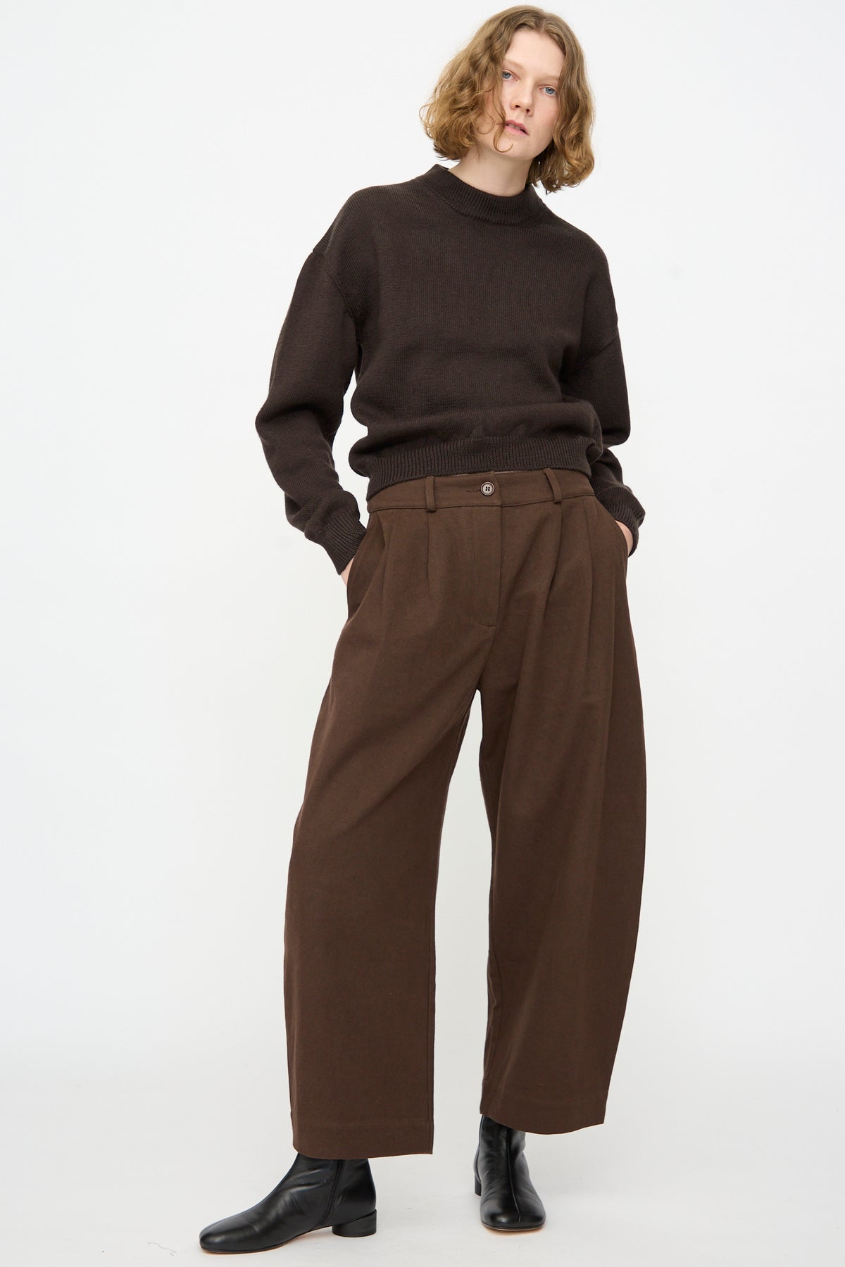 A person wearing a dark brown sweater, Cawley Japanese Gabardine Robyn Trouser in Espresso, and black ankle boots stands against a plain white background with hands in pockets.