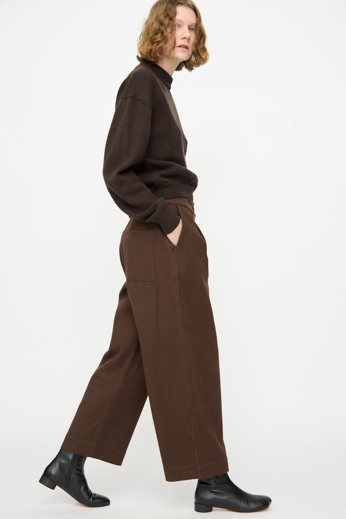 A person wearing a dark brown sweater, the Cawley Japanese Gabardine Robyn Trouser in Espresso featuring a relaxed fit and balloon leg silhouette, and black ankle boots, stands sideways with hands in pockets against a white background.