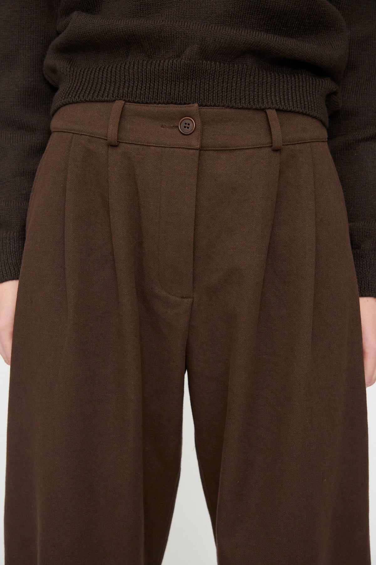 Wearing the Cawley Japanese Gabardine Robyn Trouser in Espresso—a high-waisted, relaxed fit pant with a single button and belt loops, crafted from Japanese gabardine cotton and featuring subtly balloon-shaped legs—paired with a dark knitted sweater. Only the torso and upper legs are visible.