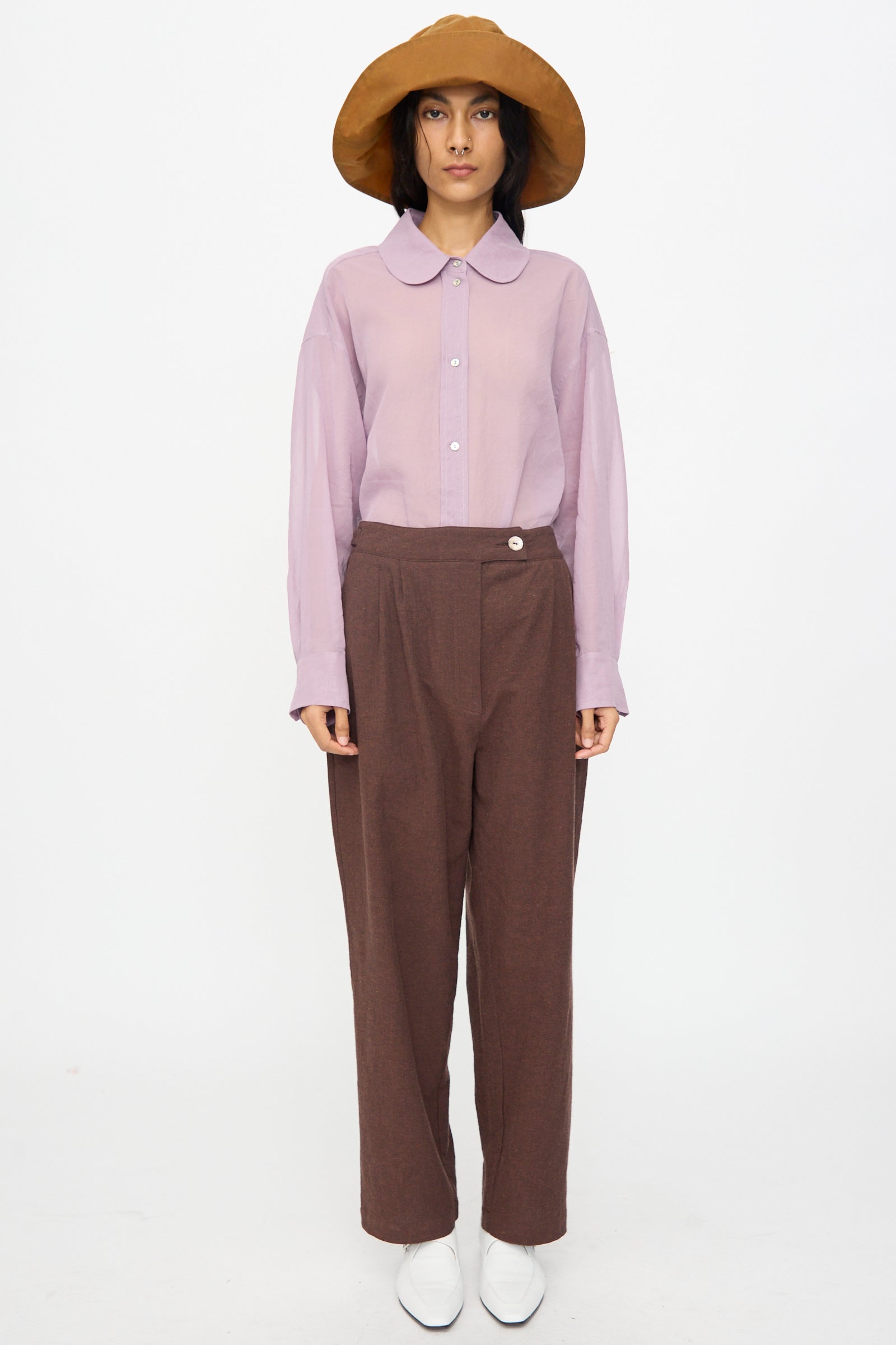 A person stands against a plain background, wearing a wide-brimmed brown hat, light purple long-sleeve shirt, Cawley Japanese High Twisted Poplin Enya Trouser in Fog Purple, and white shoes.