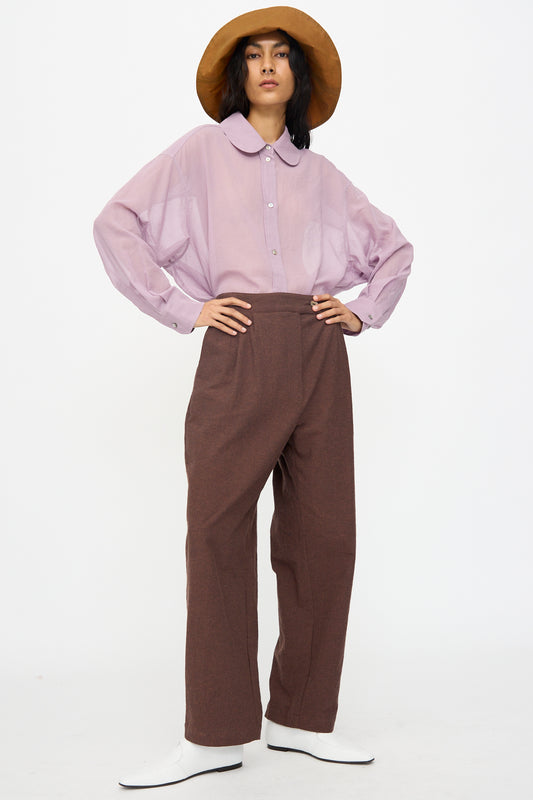 A person stands with hands on hips against a white background, wearing the Cawley Japanese High Twisted Poplin Enya Trouser in Fog Purple paired with a light purple button-up shirt and a wide-brimmed brown hat.
