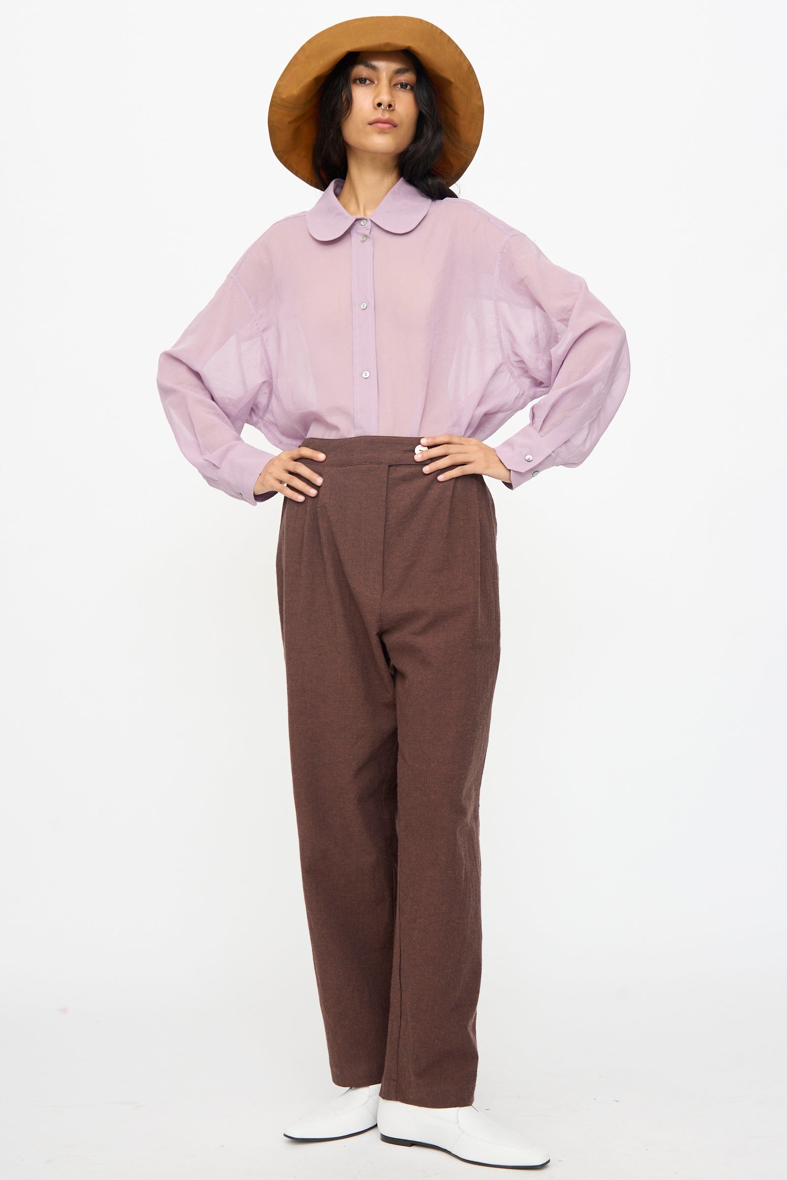 Person stands against a white background, hands on hips, wearing a wide-brimmed brown hat, light purple long-sleeve shirt, white shoes, and Cawley’s Japanese High Twisted Poplin Enya Trouser in Fog Purple with an adjustable waistband.