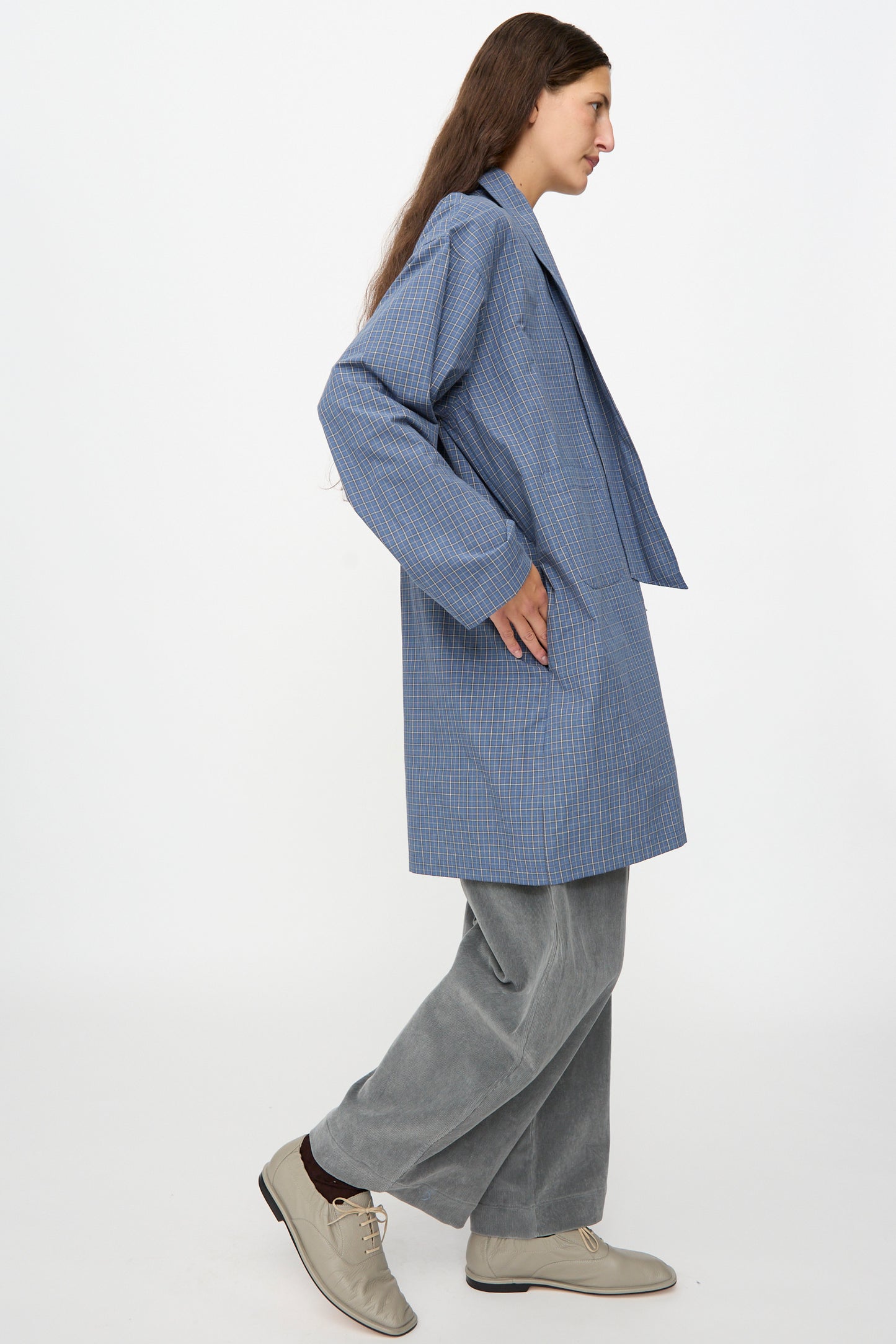 A person wearing the Cawley Japanese Mini Check Cotton Dorothy Dress in Blue walks to the left against a plain white background, styled with gray wide-leg pants and beige shoes.