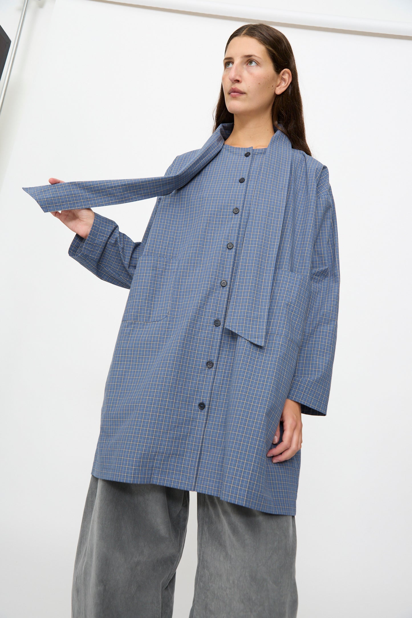 A woman wearing the Cawley Japanese Mini Check Cotton Dorothy Dress in Blue stands against a white background, styled with a matching scarf and wide gray pants.