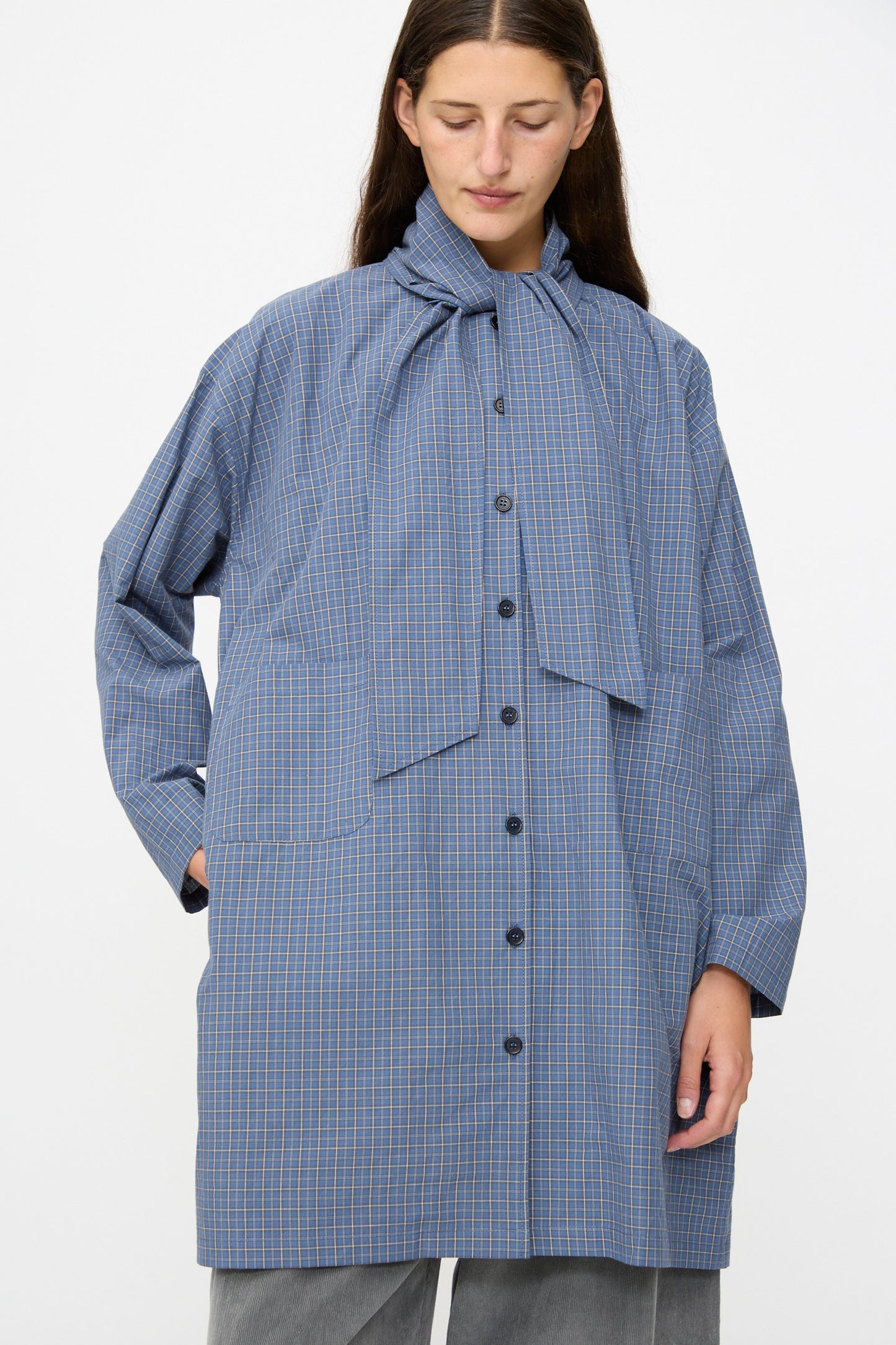 A person wearing the Cawley Japanese Mini Check Cotton Dorothy Dress in Blue, a loose-fitting, long-sleeve dress featuring large front pockets and a tied neck detail, crafted from Japanese check cotton and made in the UK.