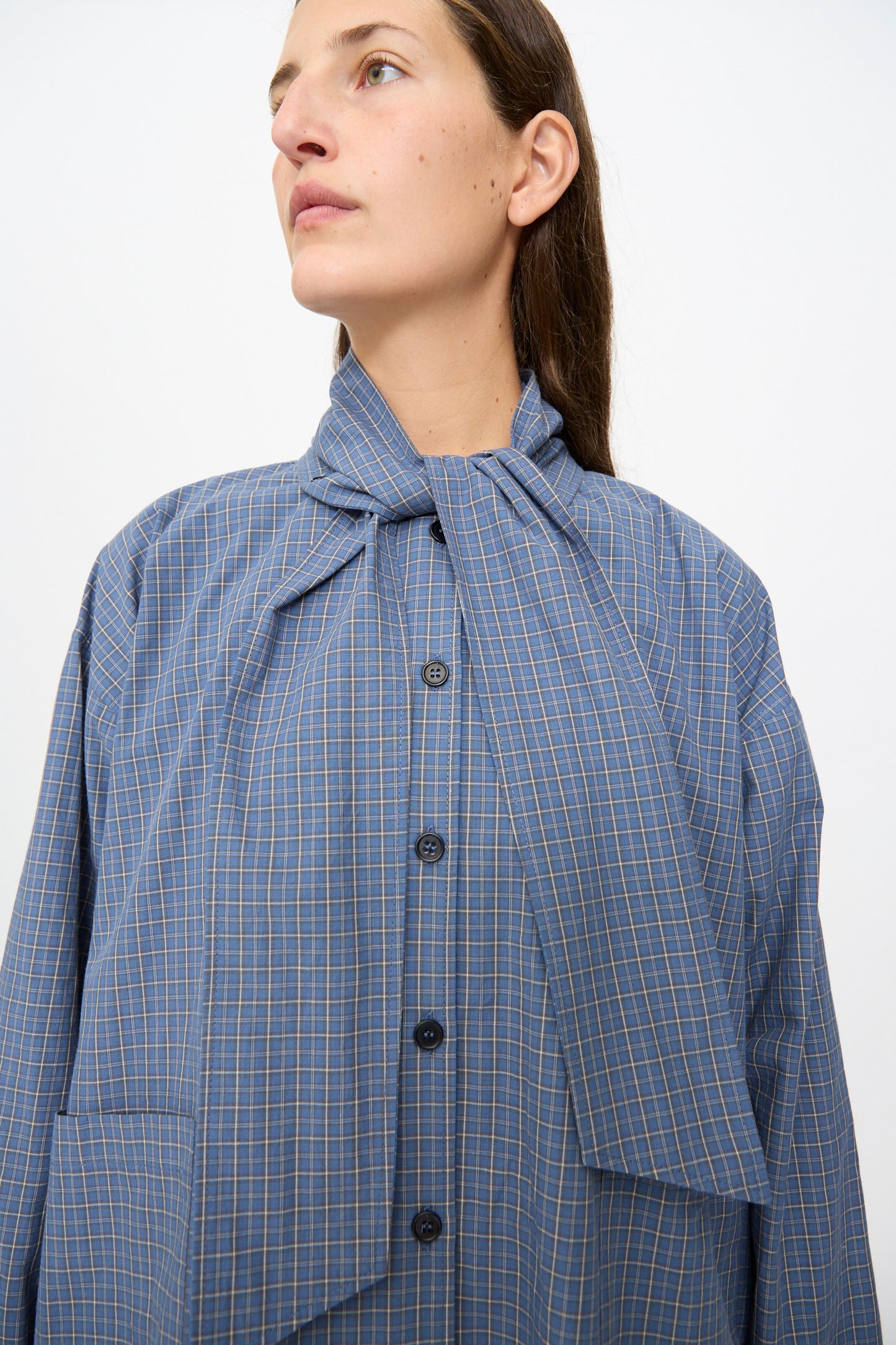 A woman wearing the Cawley Japanese Mini Check Cotton Dorothy Dress in Blue, featuring a wide necktie detail, faces slightly upward against a plain background. Made in the UK.