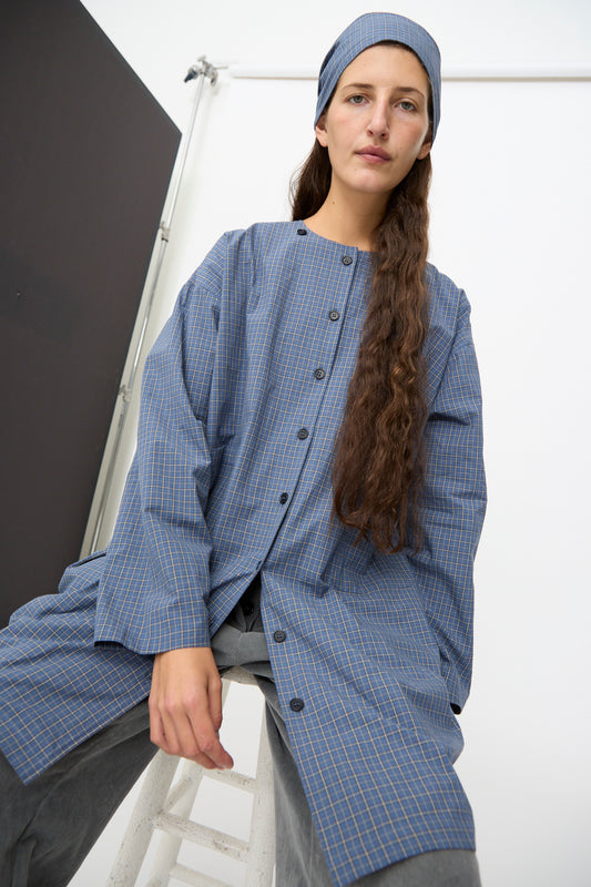 A person with long brown hair wears the Cawley Japanese Mini Check Cotton Dorothy Dress in Blue along with a matching headscarf, sitting on a stool against a minimalist studio backdrop.
