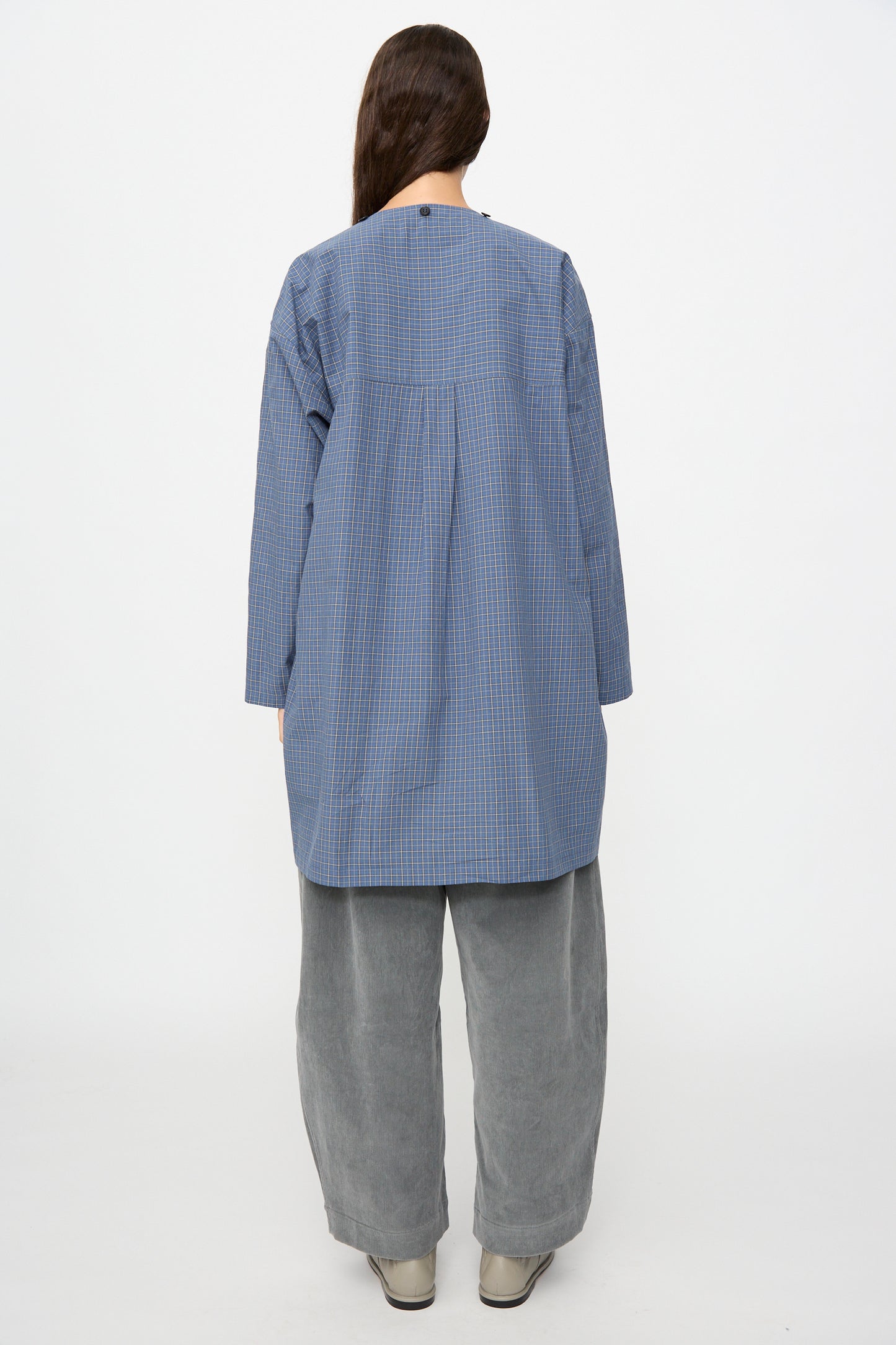 A person stands with their back to the camera, wearing the Cawley Japanese Mini Check Cotton Dorothy Dress in Blue, paired with loose grey pants and light shoes, against a plain white background.