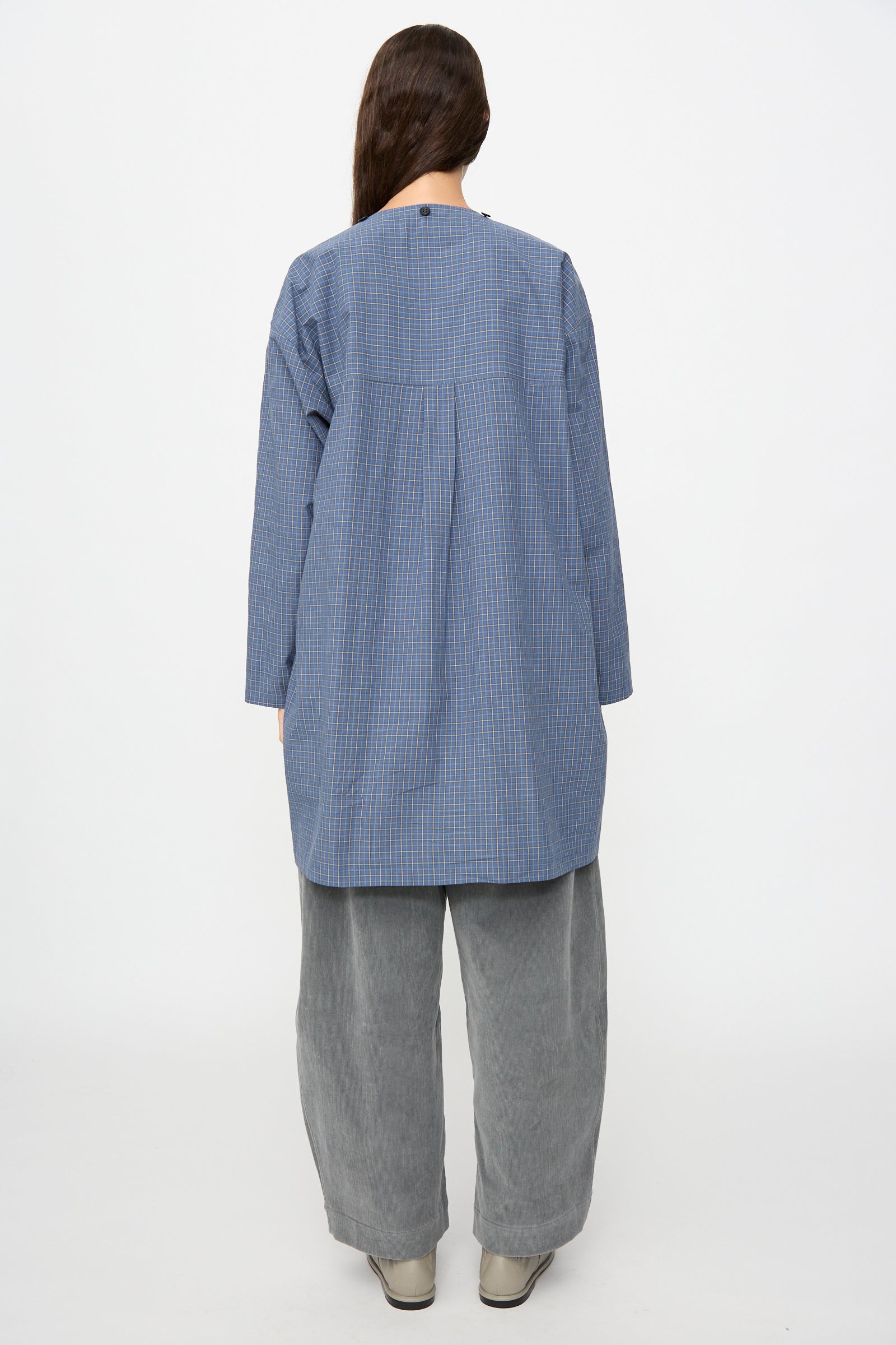 A person stands with their back to the camera, wearing the Cawley Japanese Mini Check Cotton Dorothy Dress in Blue, paired with loose grey pants and light shoes, against a plain white background.