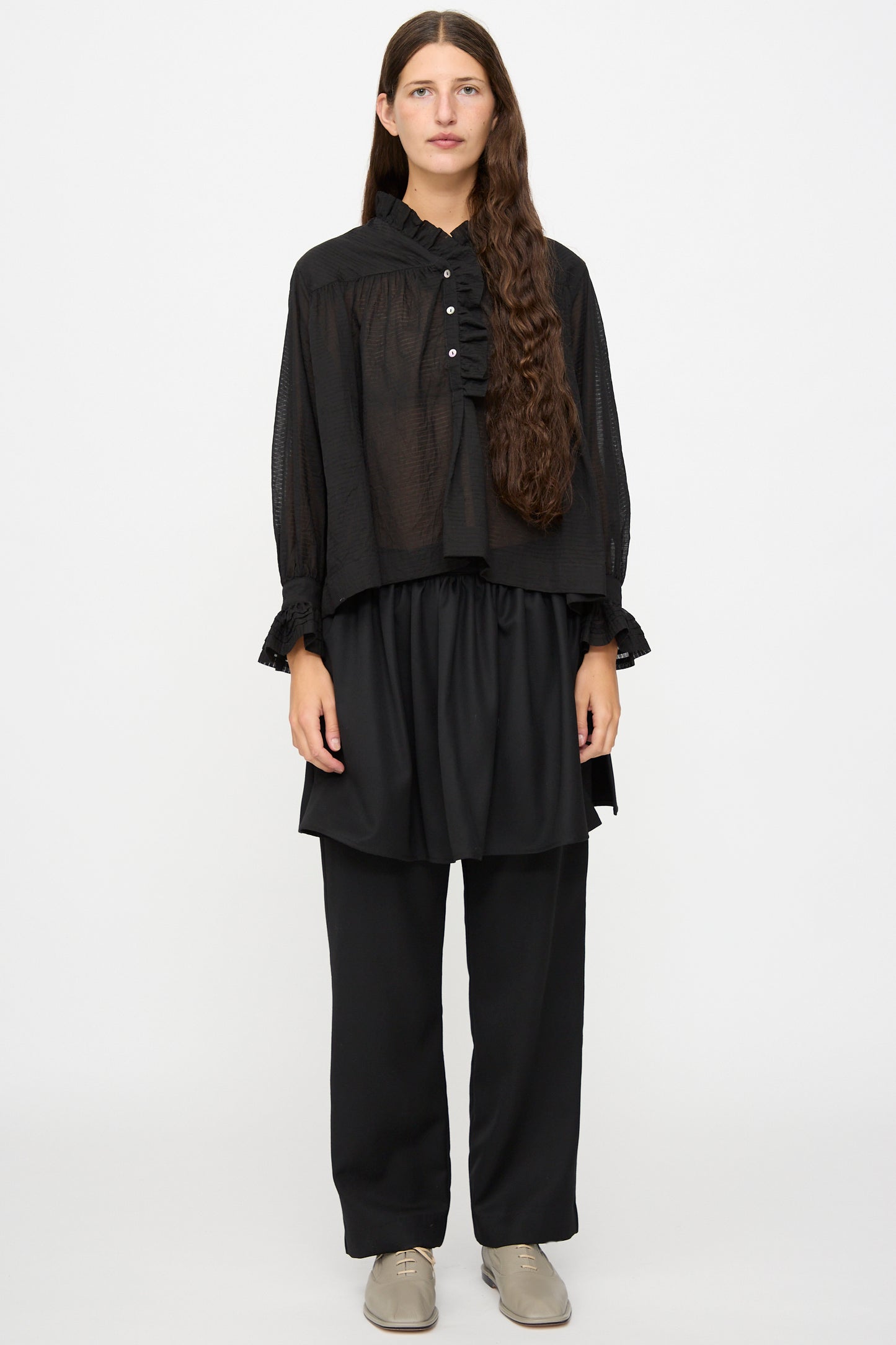 A person with long brown hair wears the Cawley Japanese Sheer Stripe Cotton Sophie Shirt in Midnight, styled as a loose black blouse, paired with a layered black skirt, black pants, and beige shoes, standing against a plain white background.