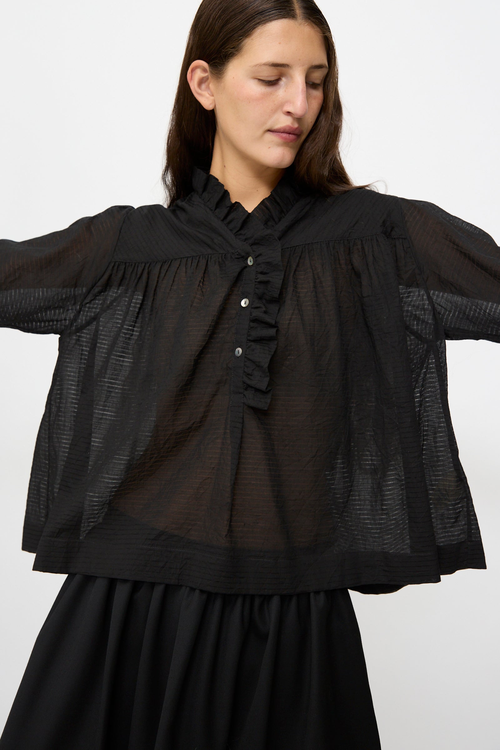 A woman wearing the Cawley Japanese Sheer Stripe Cotton Sophie Shirt in Midnight, featuring a ruffle collar and front button placket, paired with a black skirt, stands against a plain white background.